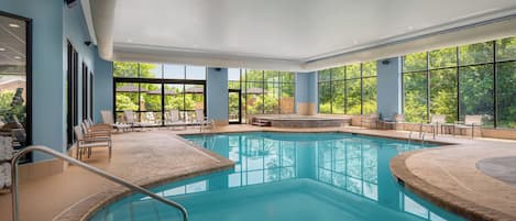 An indoor swimming pool with clear blue water, surrounded by lounge chairs and a view of greenery outside.