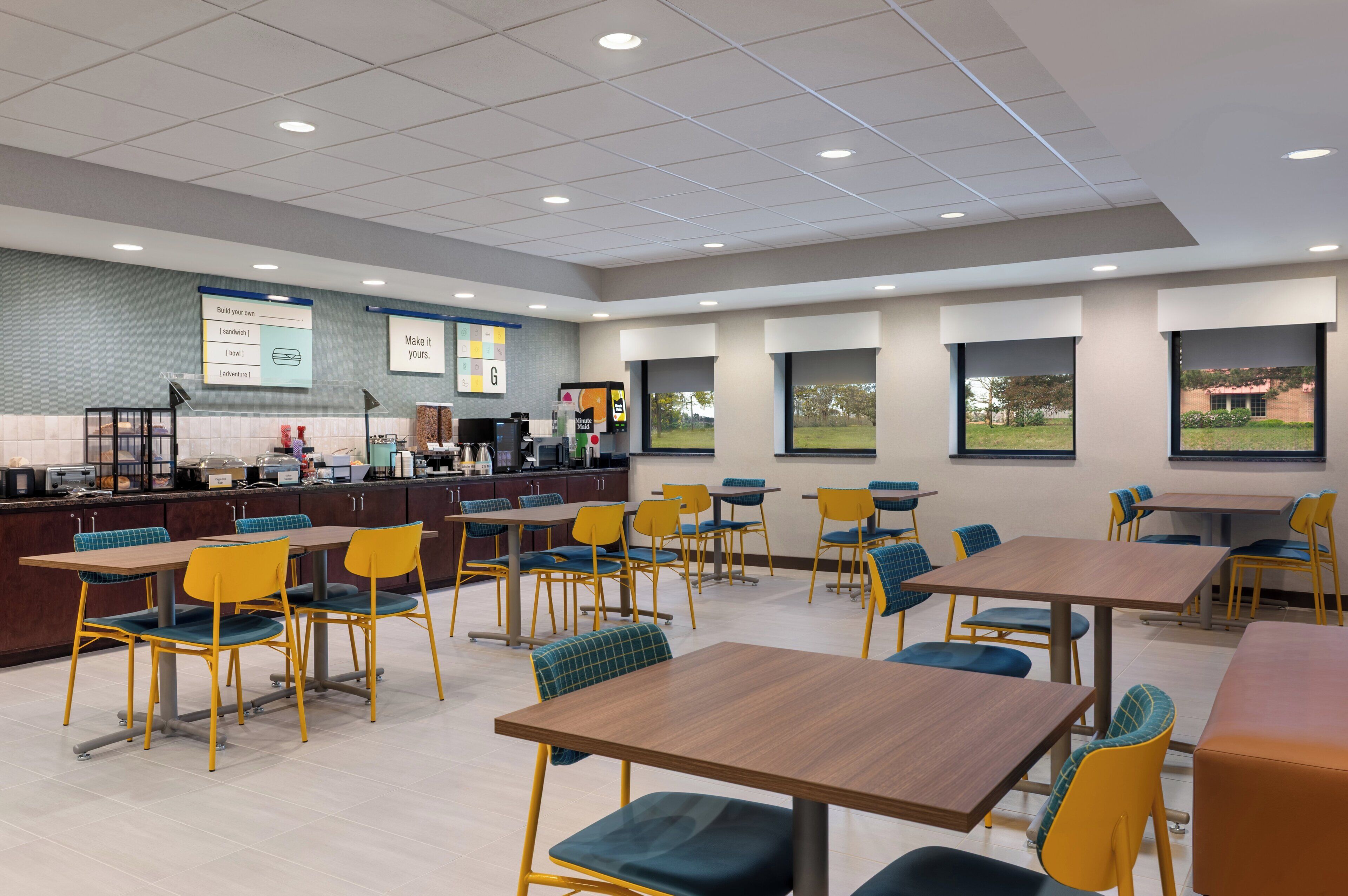 A modern cafeteria with multiple tables and chairs, a counter with food service equipment, and large windows offering views of the outdoors.