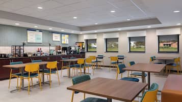 A modern cafeteria with multiple tables and chairs, a counter with food service equipment, and large windows offering views of the outdoors.