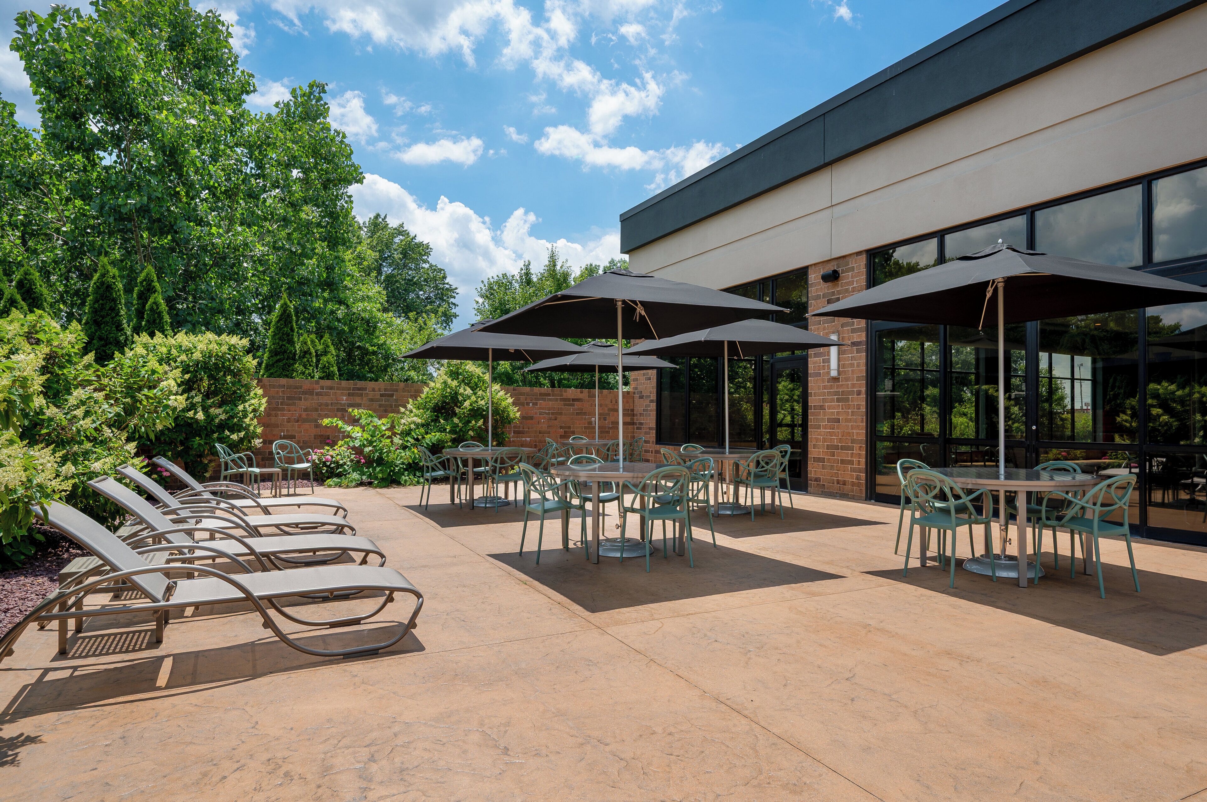 An outdoor patio with tables and chairs, surrounded by greenery and a building with large windows.