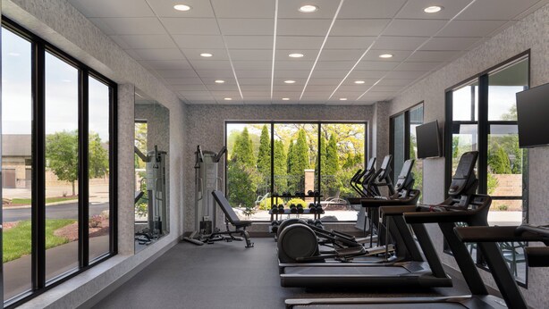 A gym with treadmills, weights, and exercise equipment, large windows, and a view of trees and a building.
