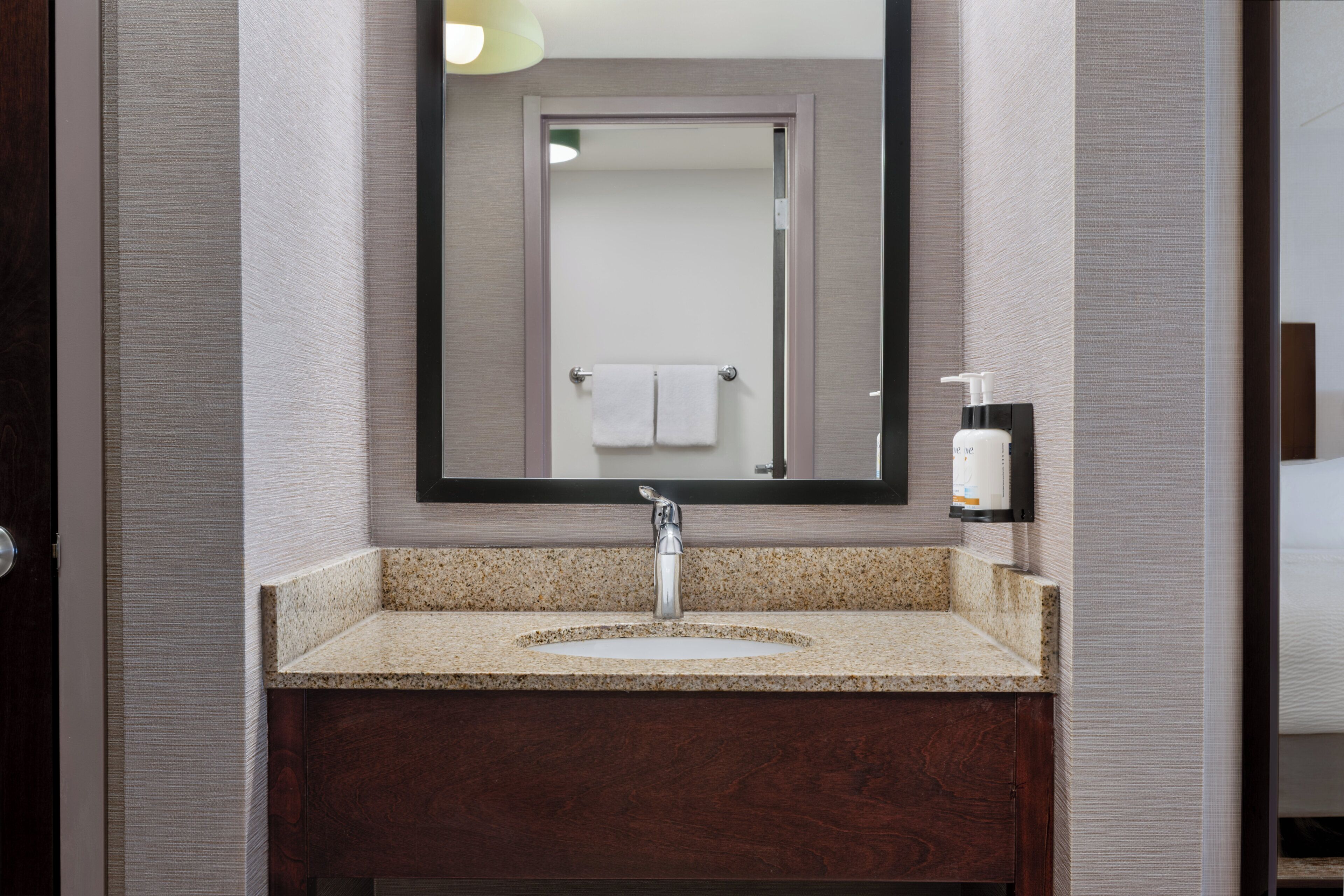 A bathroom with a granite countertop sink, a mirror, a towel rack, and a soap dispenser.
