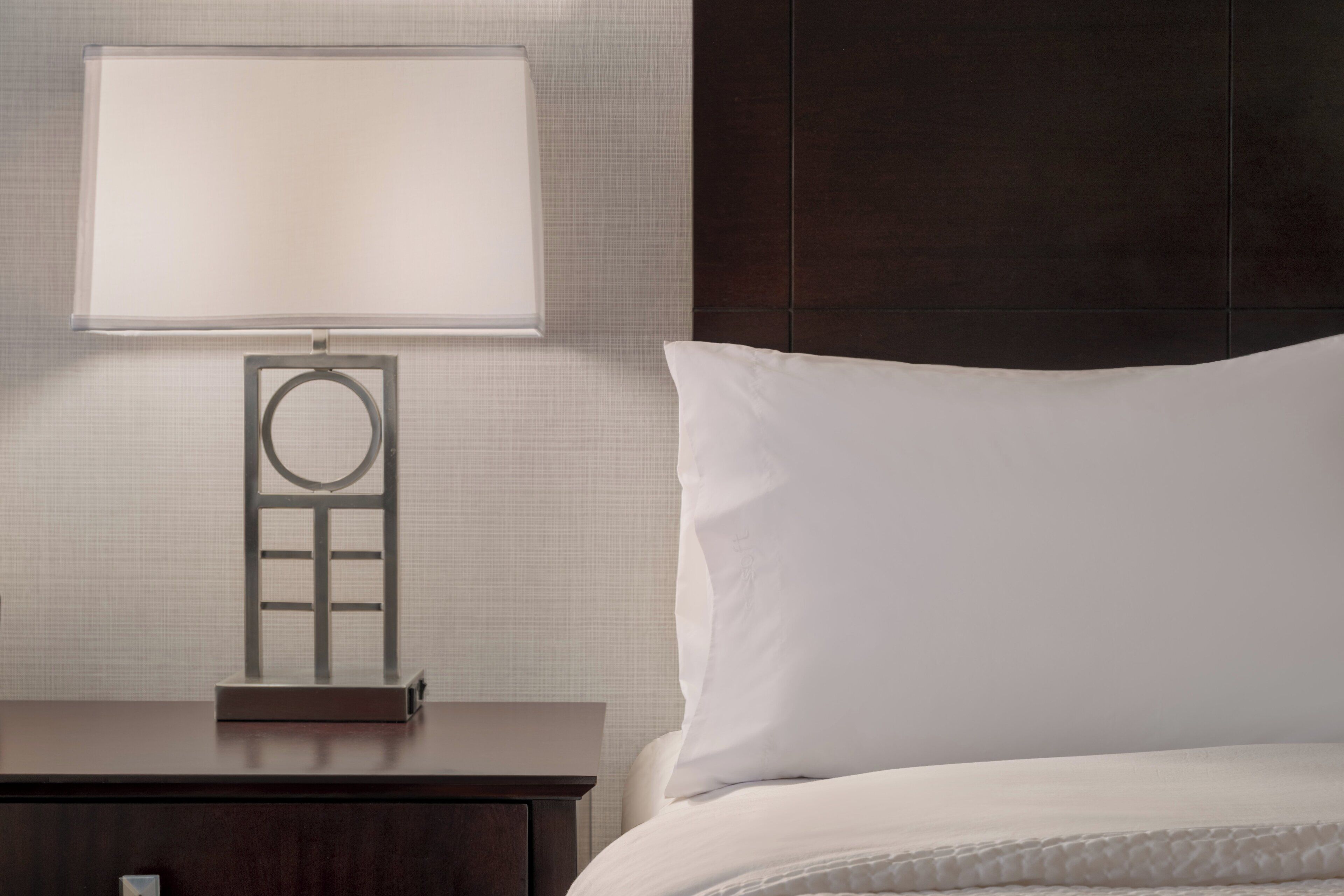 A neatly made bed with a white pillow and a dark wooden nightstand with a lamp.