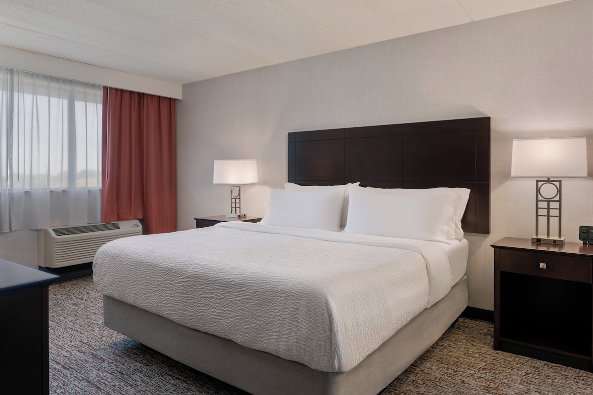 Une chambre d’hôtel avec un grand lit, deux tables de chevet avec des lampes, une fenêtre avec des rideaux et un appareil de climatisation.