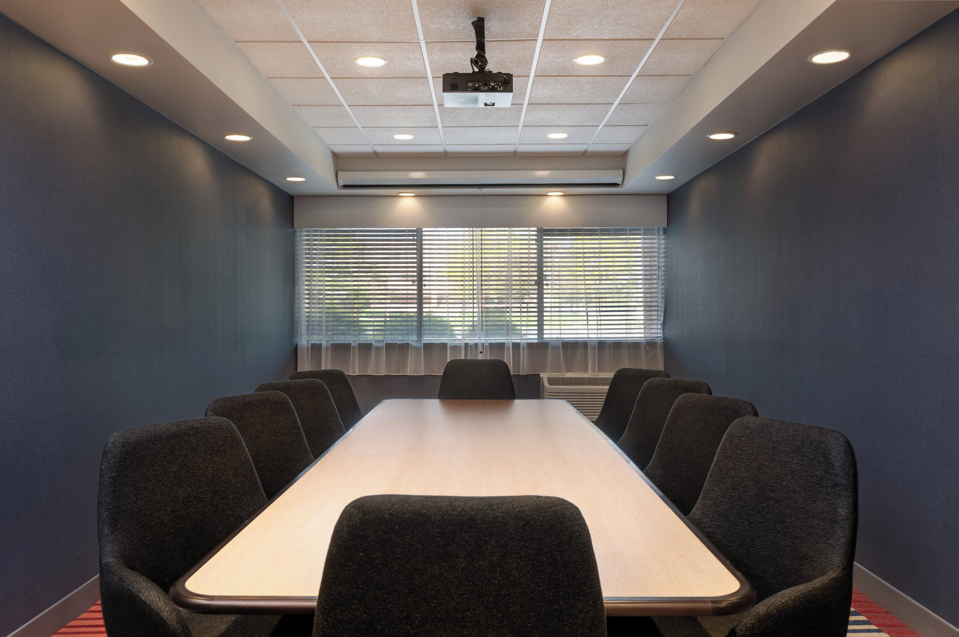 A conference room with a long table, six chairs, ceiling lights, and windows with blinds.