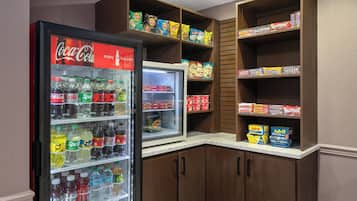 Un refrigerador lleno de bebidas y aperitivos, un microondas y estantes con alimentos enlatados.