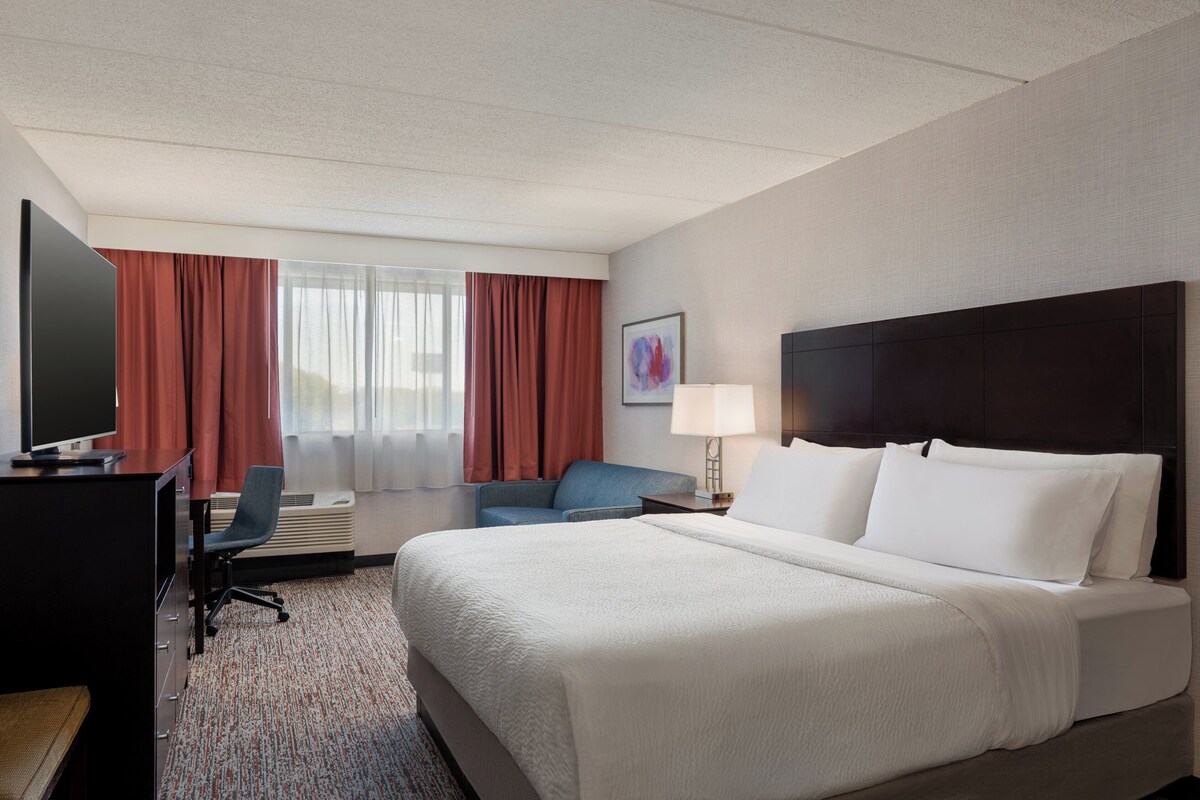 Une chambre d’hôtel avec un lit, un bureau avec une télévision, une chaise, une lampe et une fenêtre avec des rideaux.