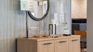 A hotel reception area with a wooden cabinet, a glass water dispenser, a mirror, and a sign with amenities.