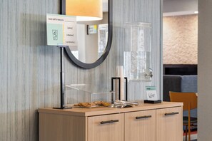 Un'area reception di un hotel con un mobiletto in legno, un distributore d'acqua in vetro, uno specchio e un cartello che illustra i servizi.