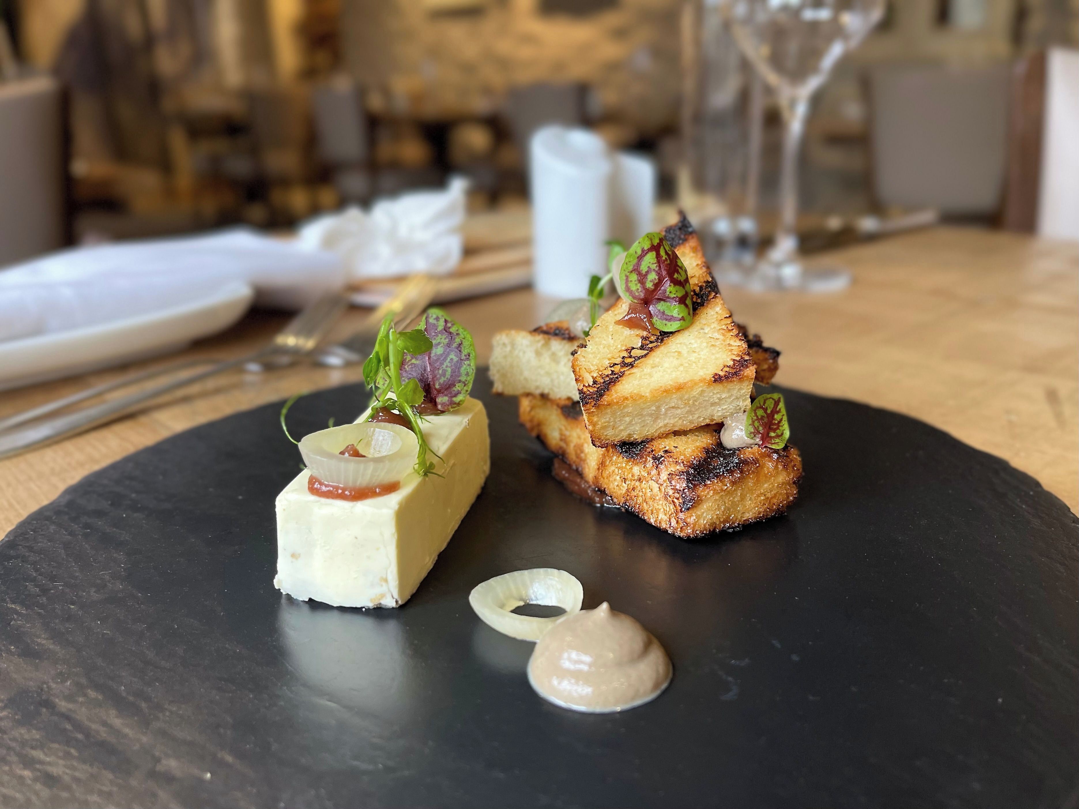 A black slate plate with two gourmet dishes, a glass of wine, and a napkin on a wooden table.