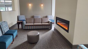 A modern lounge area with a sofa, ottoman, and patterned chairs. A fireplace with a glass front is on the wall.