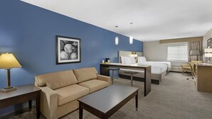 Une chambre d’hôtel avec un mur d’accent bleu, un canapé beige, une table basse en bois, un bureau avec une lampe, un lit avec du linge de maison blanc et un tableau encadré au mur.