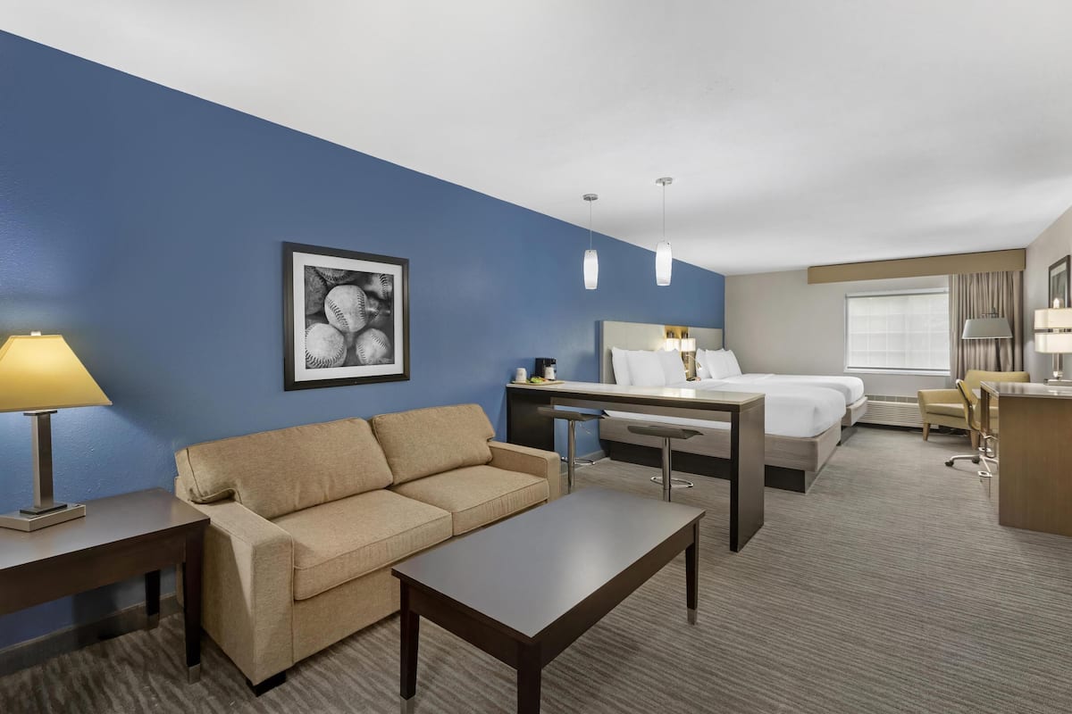 A hotel room with a blue accent wall, a beige sofa, a wooden coffee table, a desk with a lamp, a bed with white linens, and a framed picture on the wall.