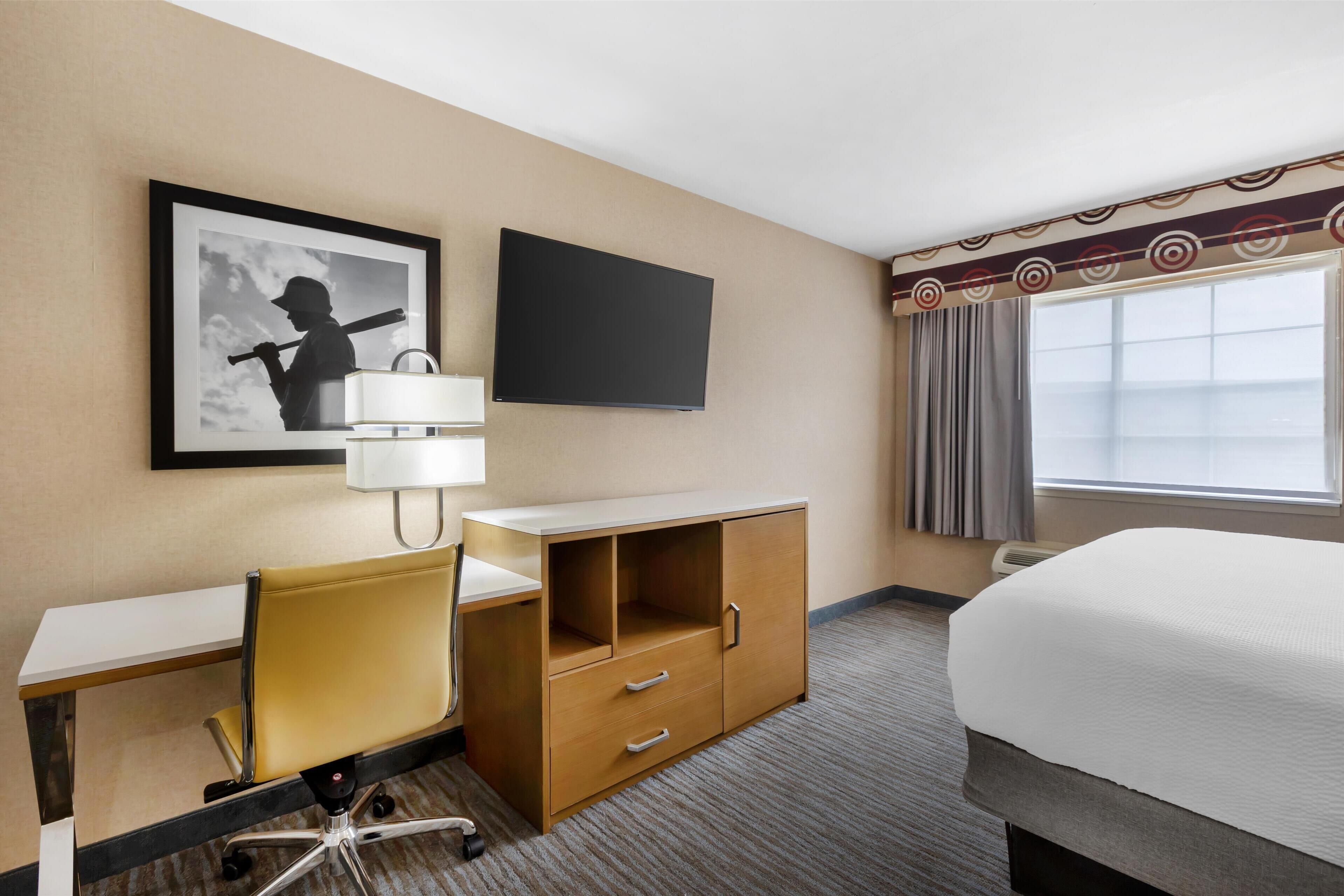 A hotel room with a bed, a desk with a chair, a TV, a framed picture on the wall, and a window with curtains.