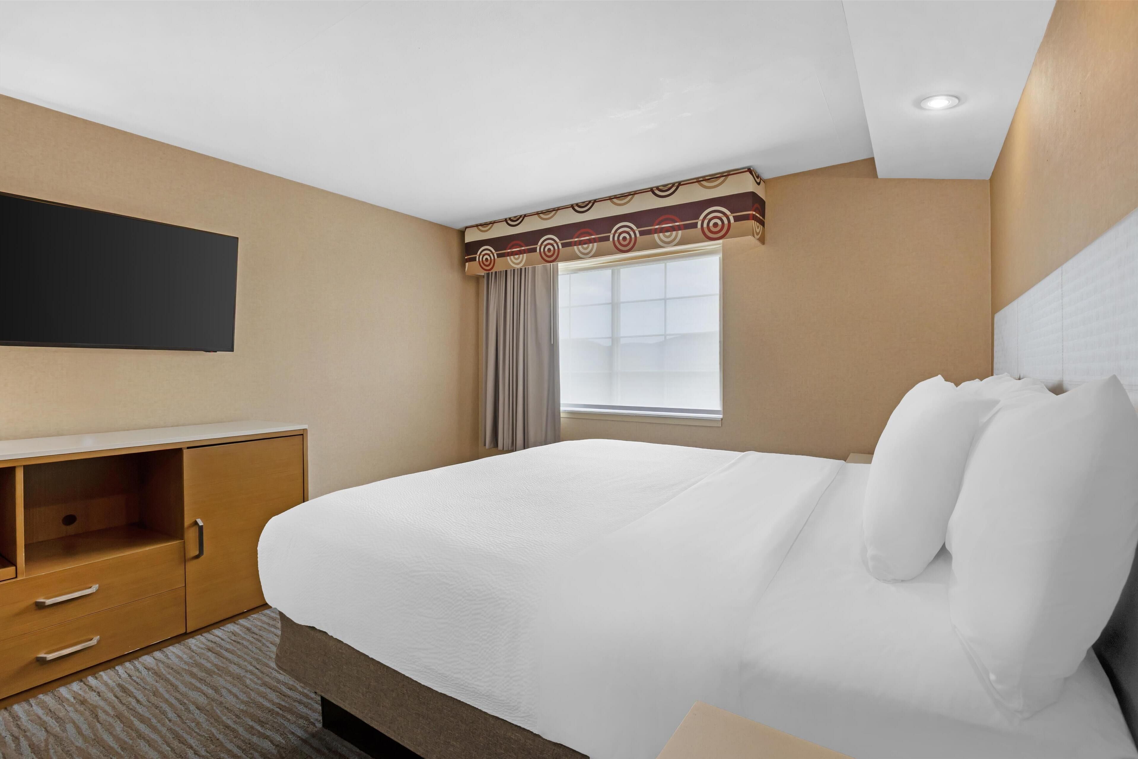 Une chambre d’hôtel dotée d’un grand lit, d’un téléviseur à écran plat fixé au mur, d’une fenêtre avec des rideaux et d’une armoire en bois avec des tiroirs.