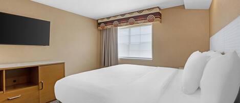 A hotel room with a large bed, a flat-screen TV mounted on the wall, a window with curtains, and a wooden cabinet with drawers.