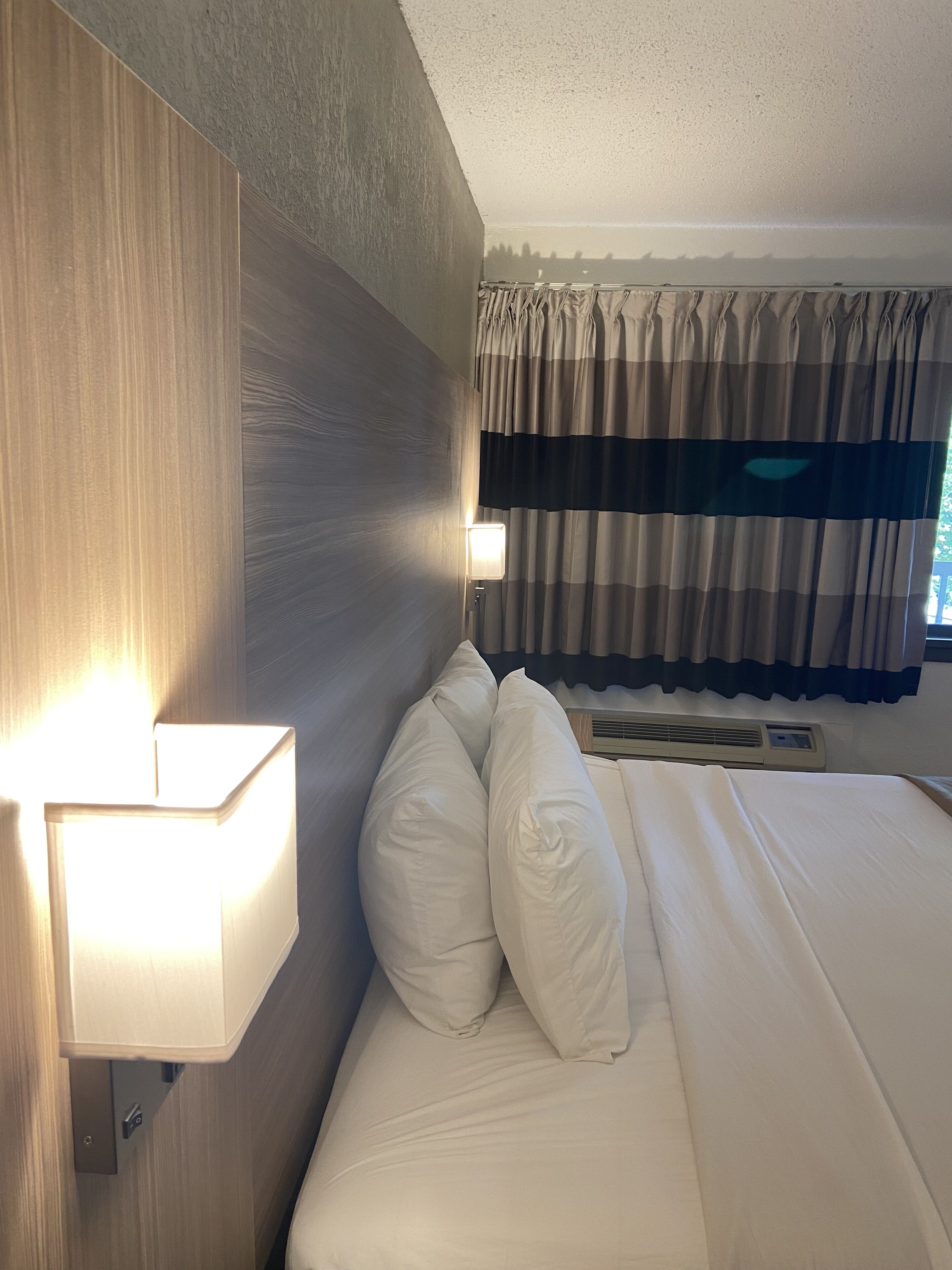 Habitación de hotel con cabecera de madera, lámpara de noche, cama con ropa blanca y ventana con cortinas a rayas.