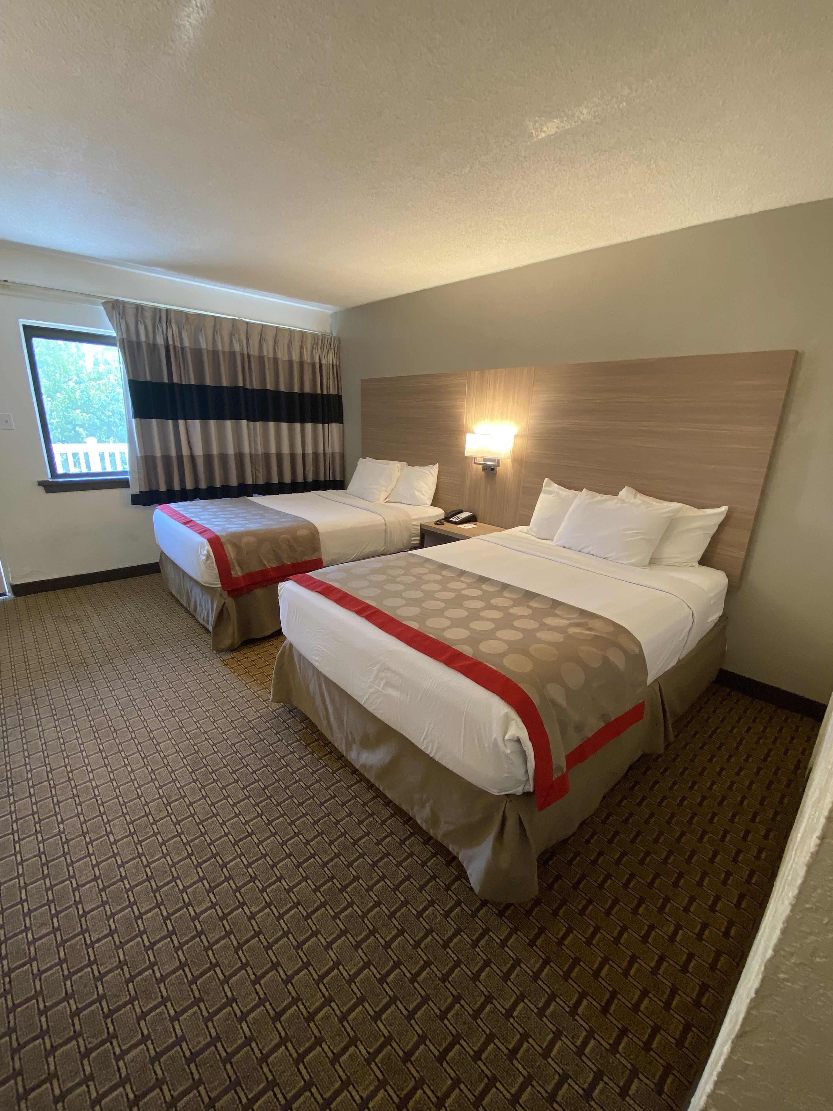 A hotel room with two beds, a window with curtains, a wall-mounted lamp, and a patterned floor.