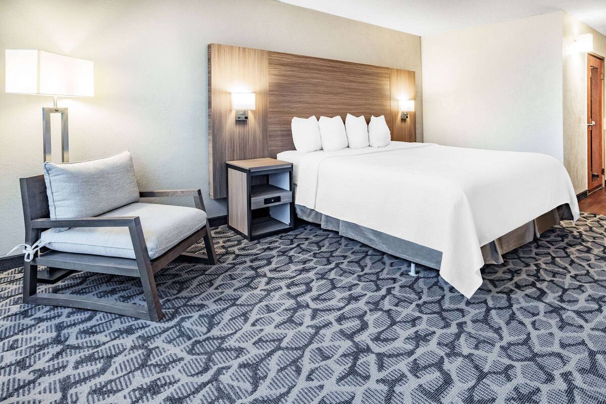 A hotel room with a bed, a chair, a nightstand, and a lamp.