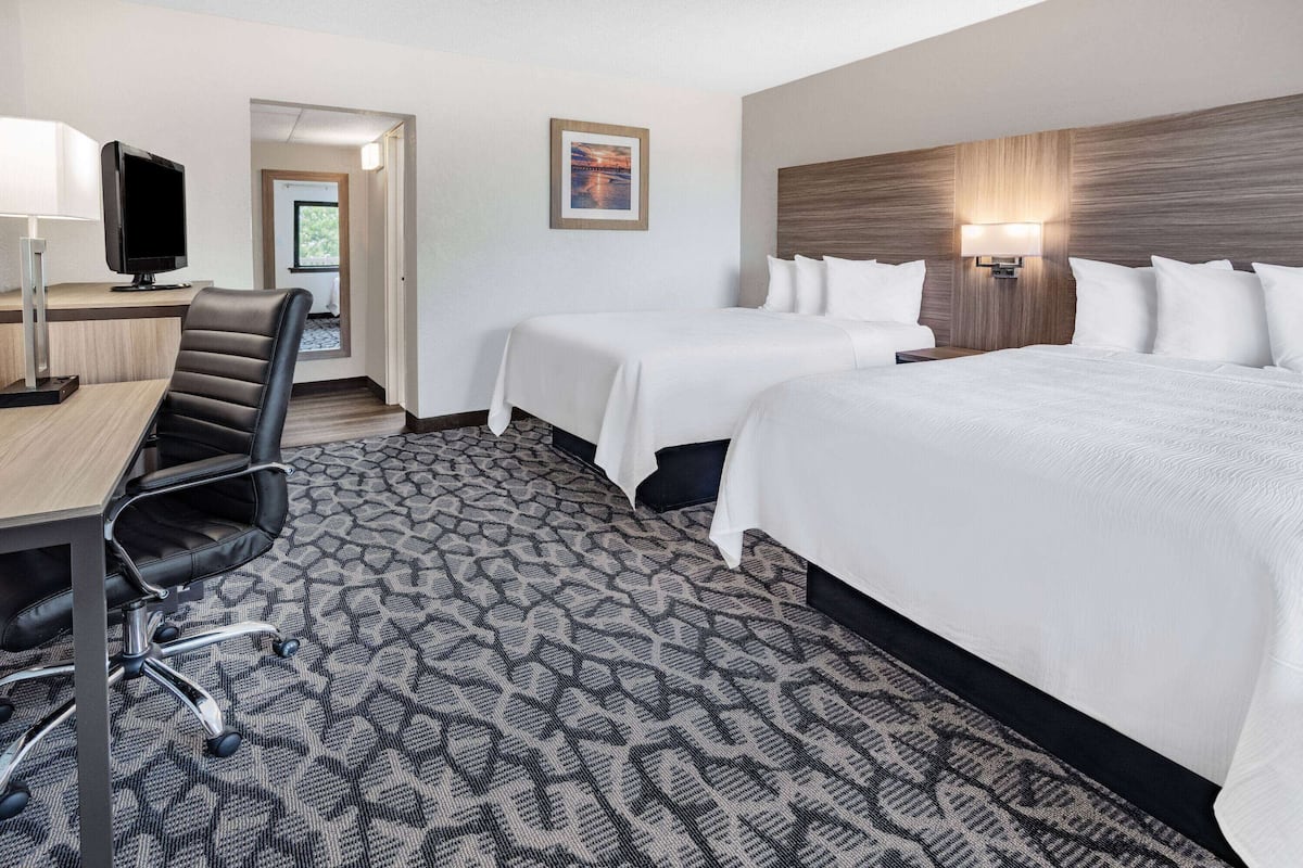 A hotel room with a desk, two beds, a chair, a TV, and a wall-mounted lamp.