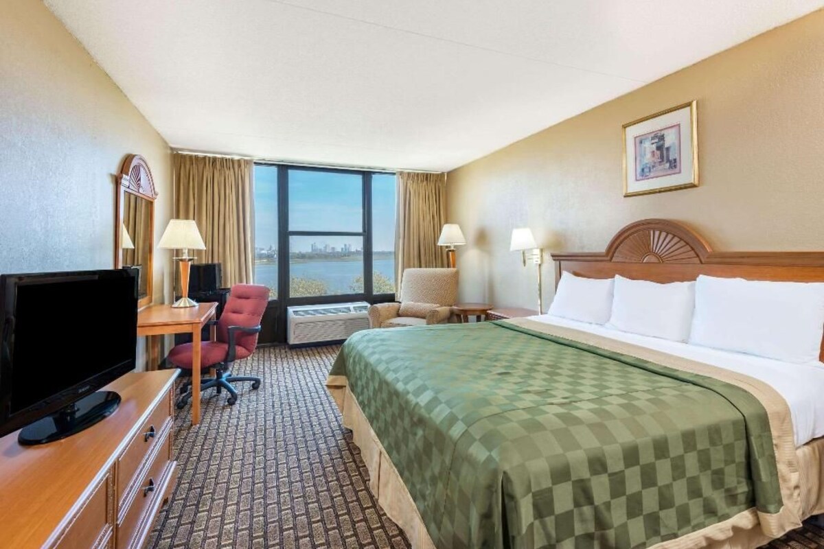 A hotel room with a bed, a desk with a television, a chair, and a view of the city through large windows.