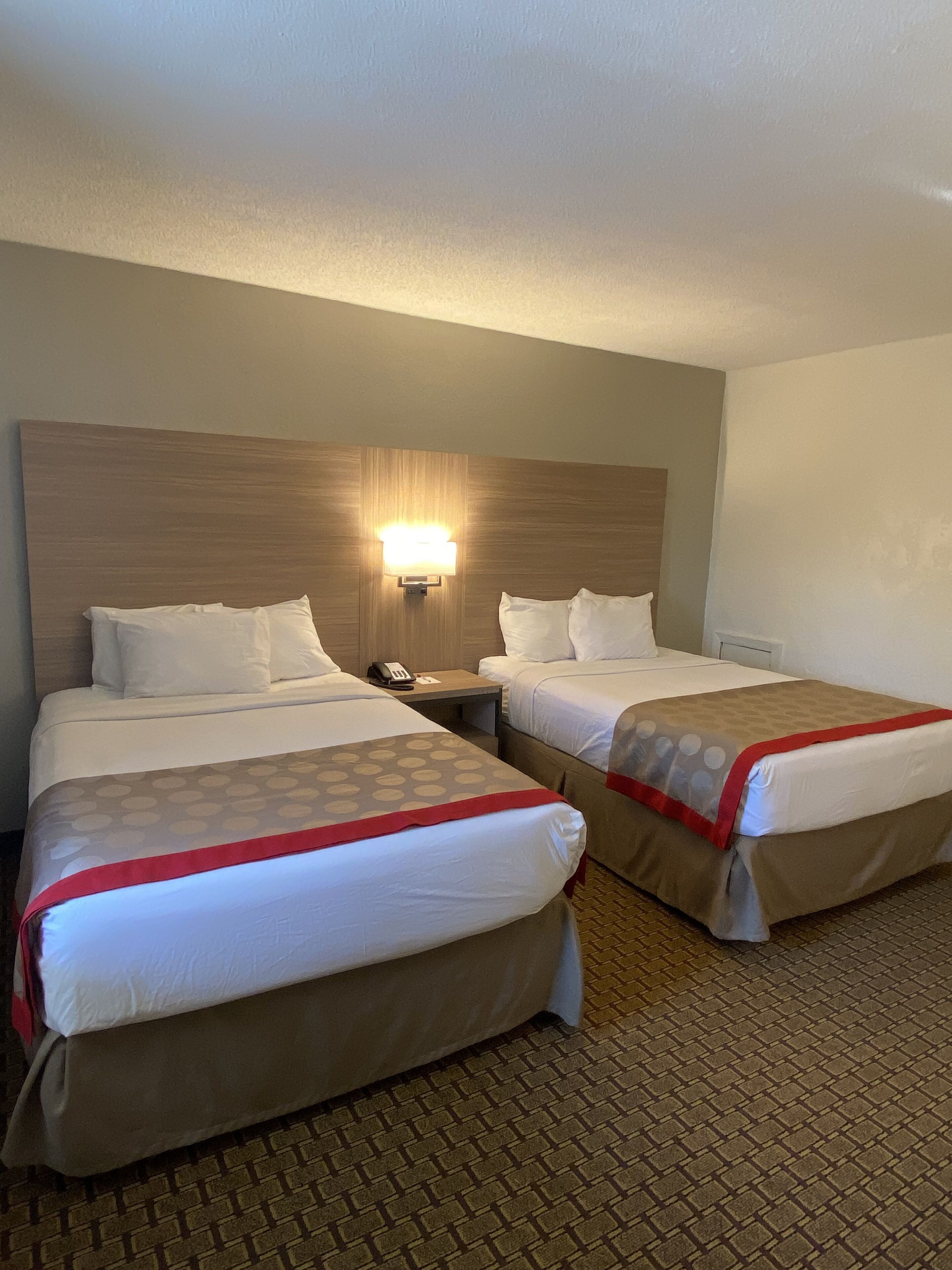 A hotel room with two beds, a nightstand with a phone, and a wall-mounted light fixture.