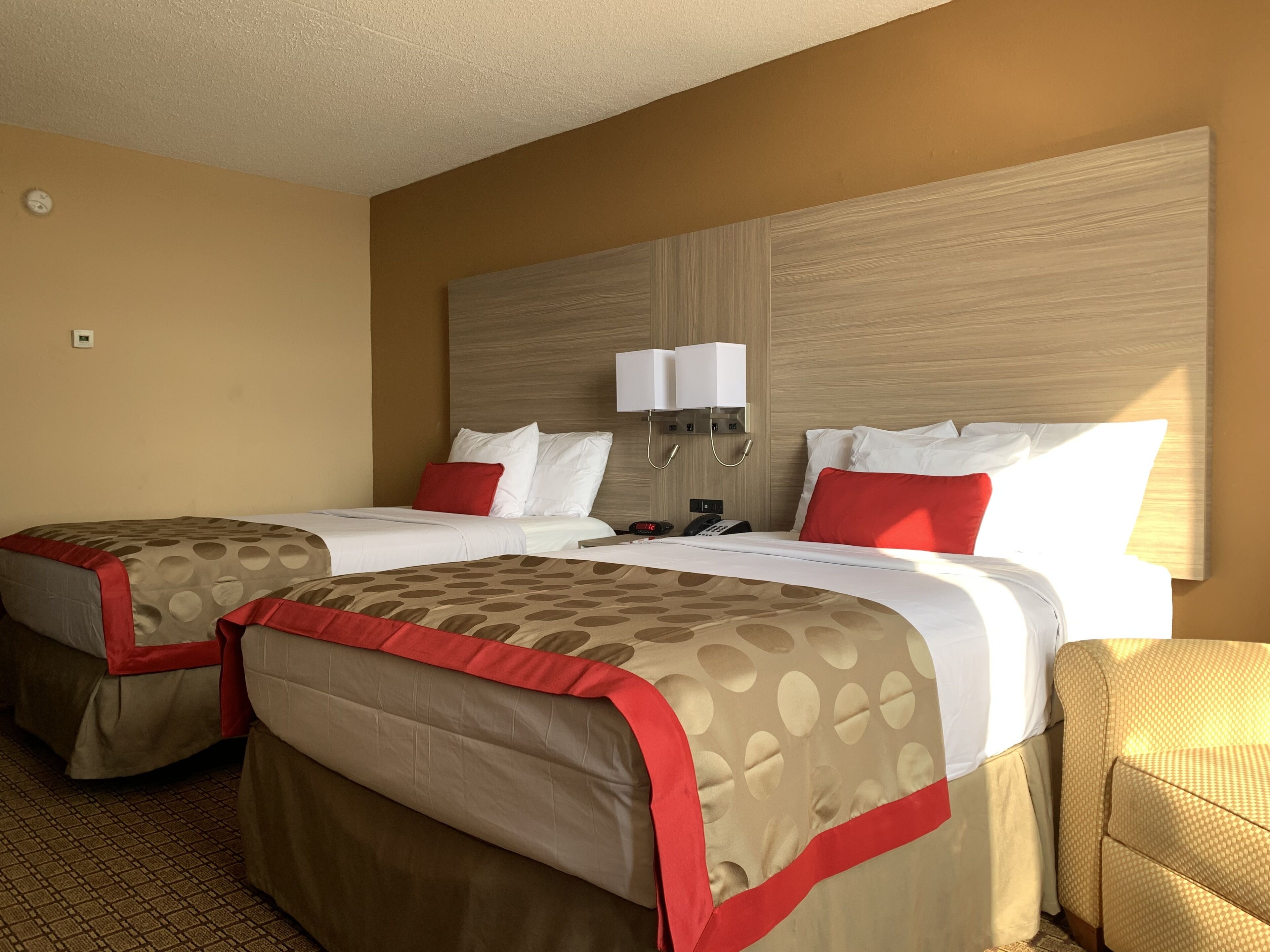 A hotel room with two beds, each with a red and beige bedspread, a wooden headboard, a bedside table with a lamp, and a wall-mounted telephone.
