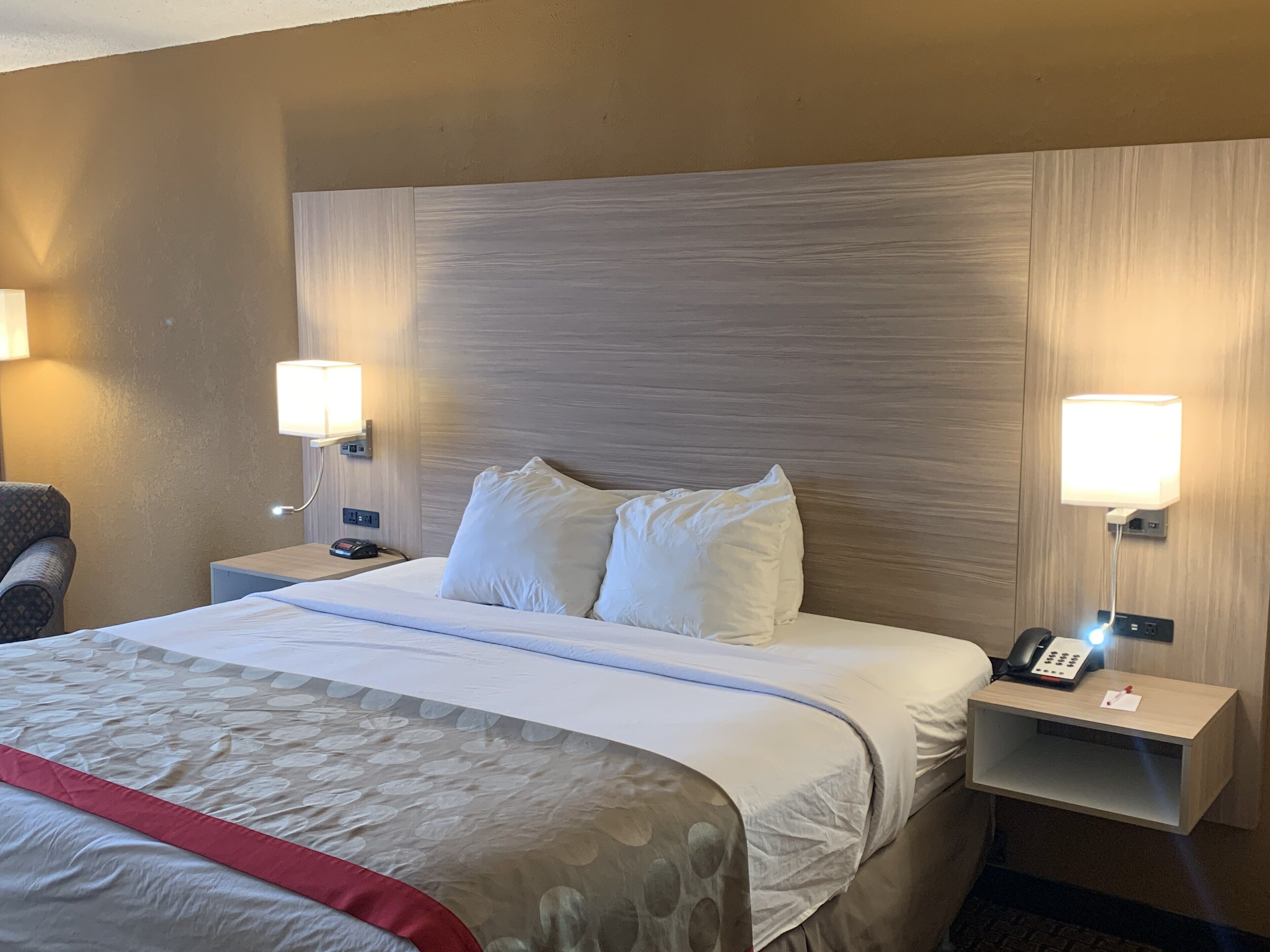 Habitación de hotel con una cama grande, dos mesitas de noche, una silla y dos lámparas de pared.