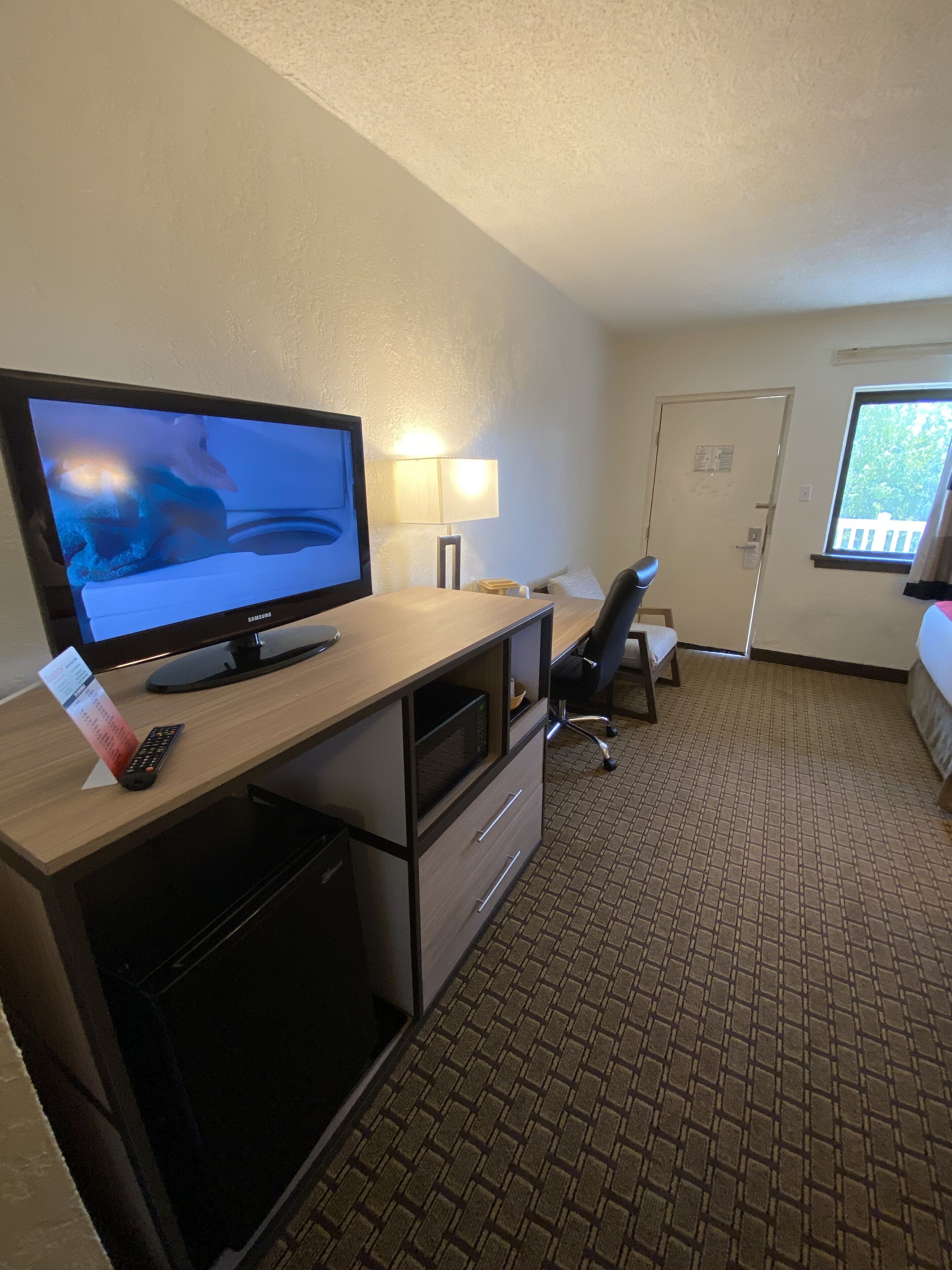 Een hotelkamer met een bureau, een televisie, een lamp, een stoel, een bed en een raam.