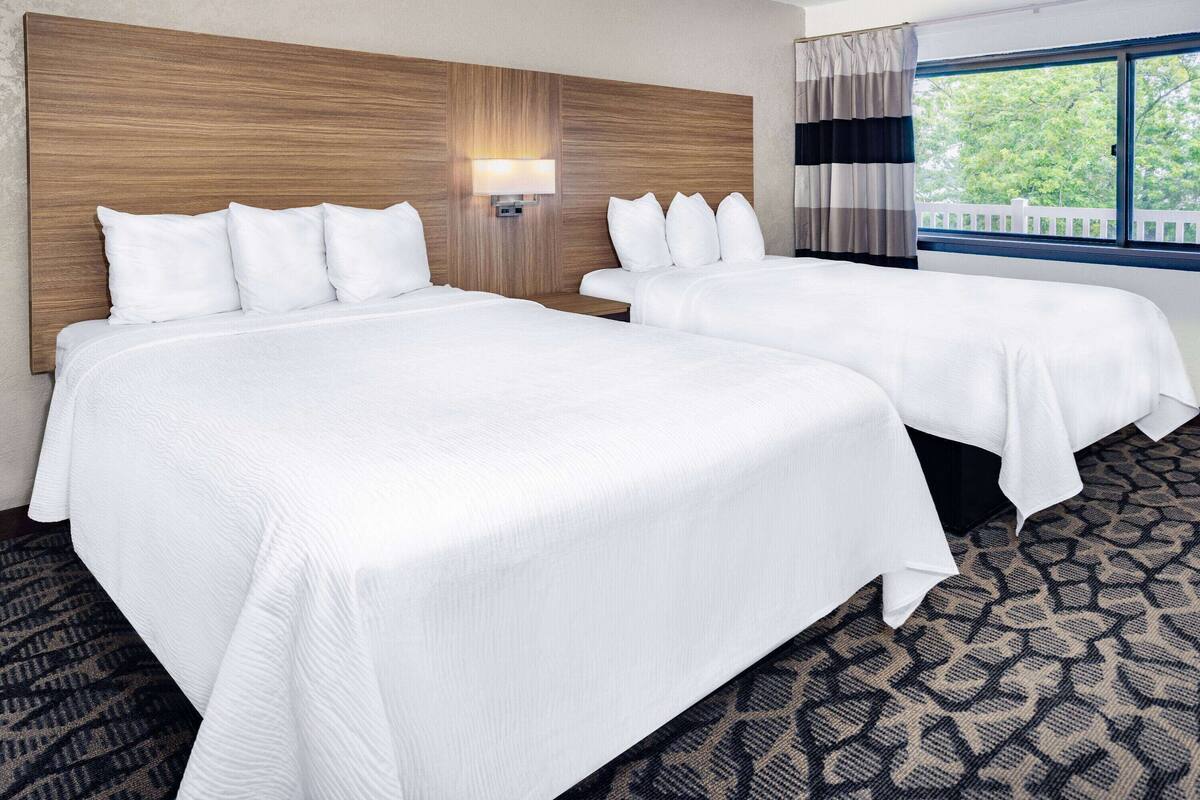 A hotel room with two beds, a wooden headboard, a nightstand with a lamp, and a window with a view of trees.