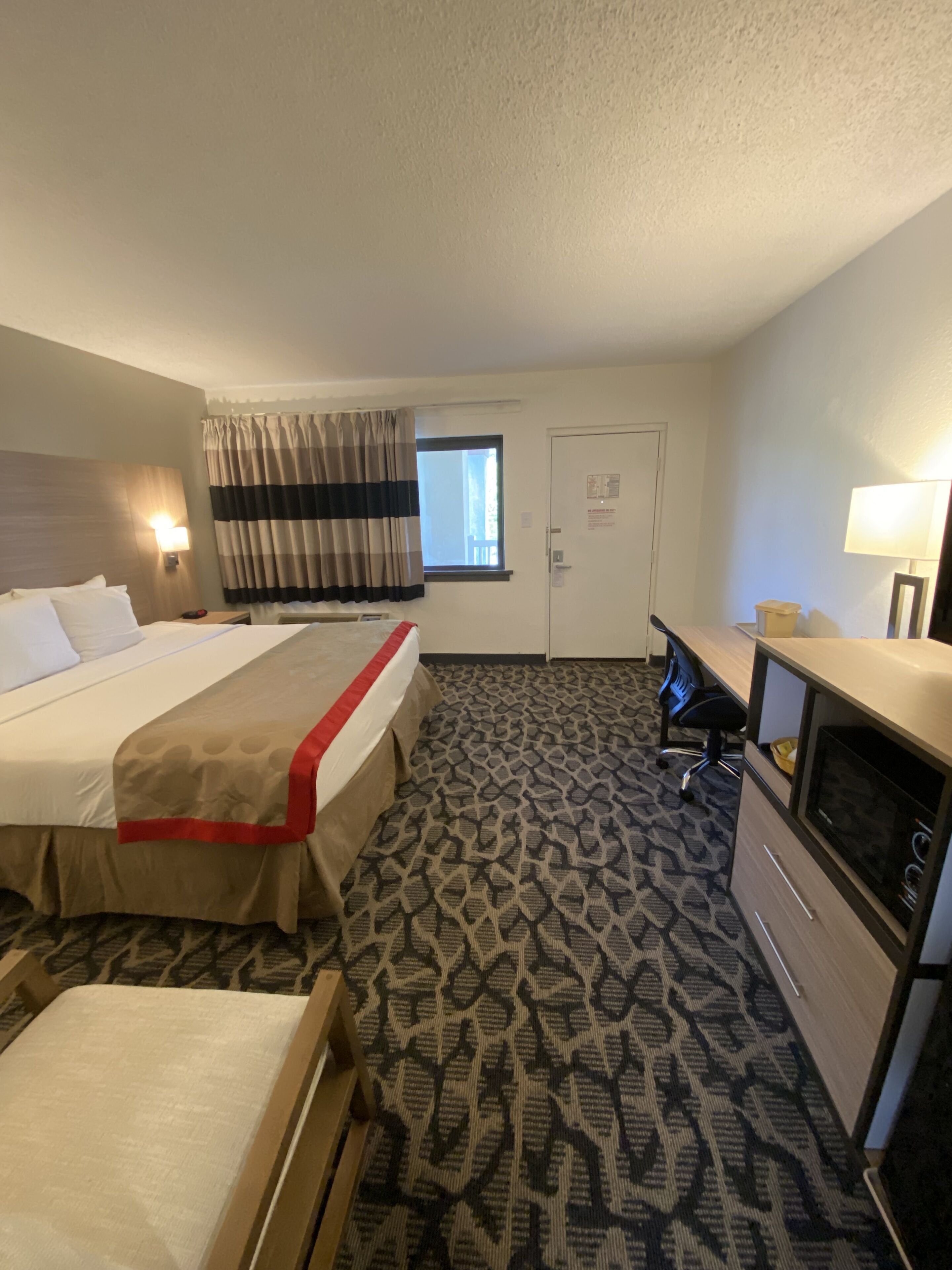 A hotel room with a large bed, a desk with a TV, a window with curtains, and a patterned carpet.
