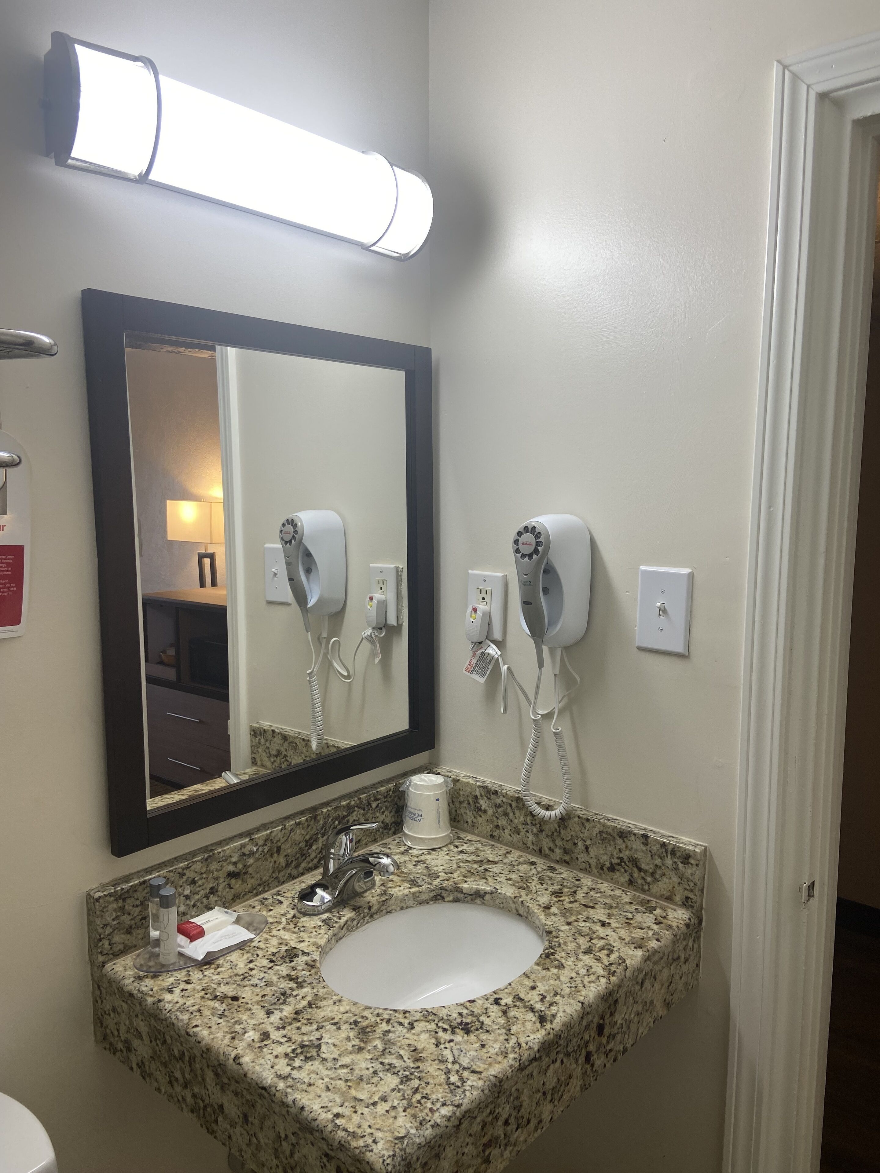 A bathroom with a granite countertop sink, a large mirror, and two hair dryers mounted on the wall.