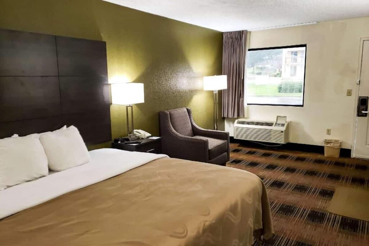 Ein Hotelzimmer mit einem Bett, einem Sessel, einem Schreibtisch mit Lampe, einem Fenster mit Vorhängen und einer Klimaanlage.