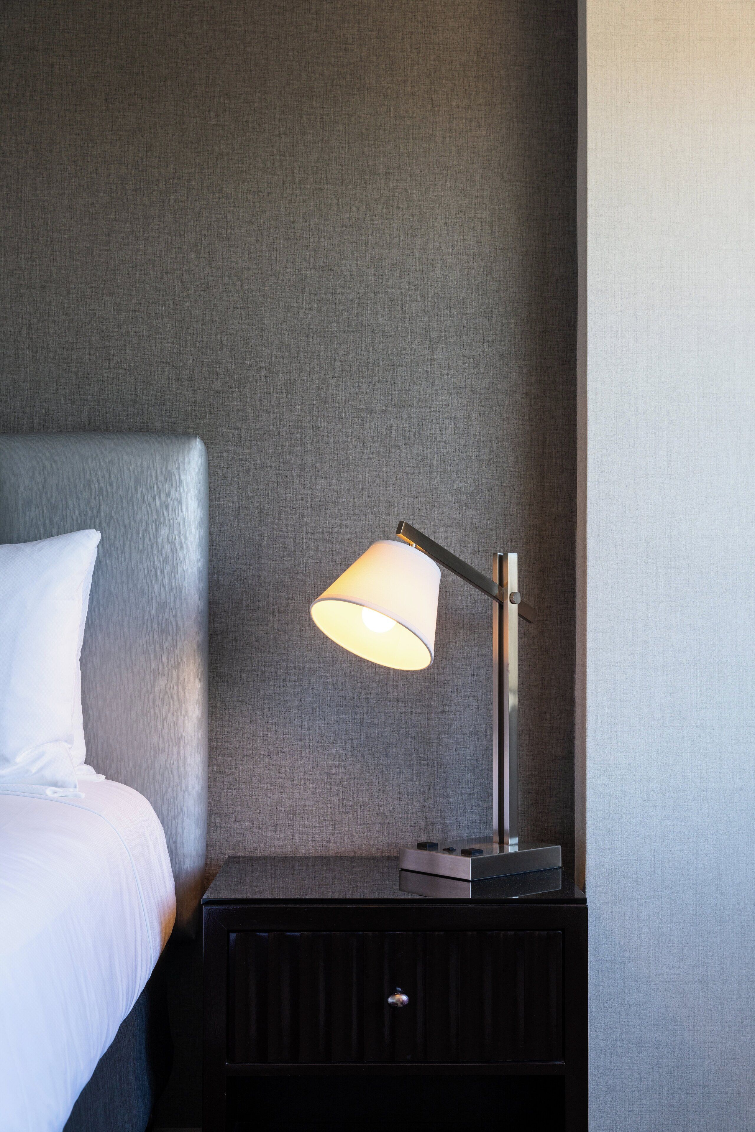 Una cama bien tendida con un cojín blanco y una mesita de noche negra a su lado. Sobre la mesita, una lámpara de mesa moderna con pantalla blanca.