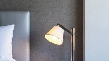 Una cama bien tendida con un cojín blanco y una mesita de noche negra a su lado. Sobre la mesita, una lámpara de mesa moderna con pantalla blanca.