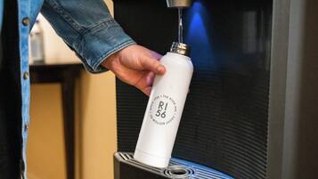 Una mano sosteniendo una botella blanca con la etiqueta 'R56' y el texto 'The River Inn' y 'No es un desagüe' en una máquina.
