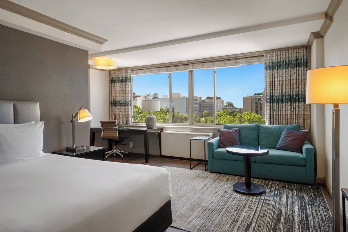 A hotel room with a large bed, a desk with a chair, a small round table, a sofa, and a view of buildings and trees through the window.