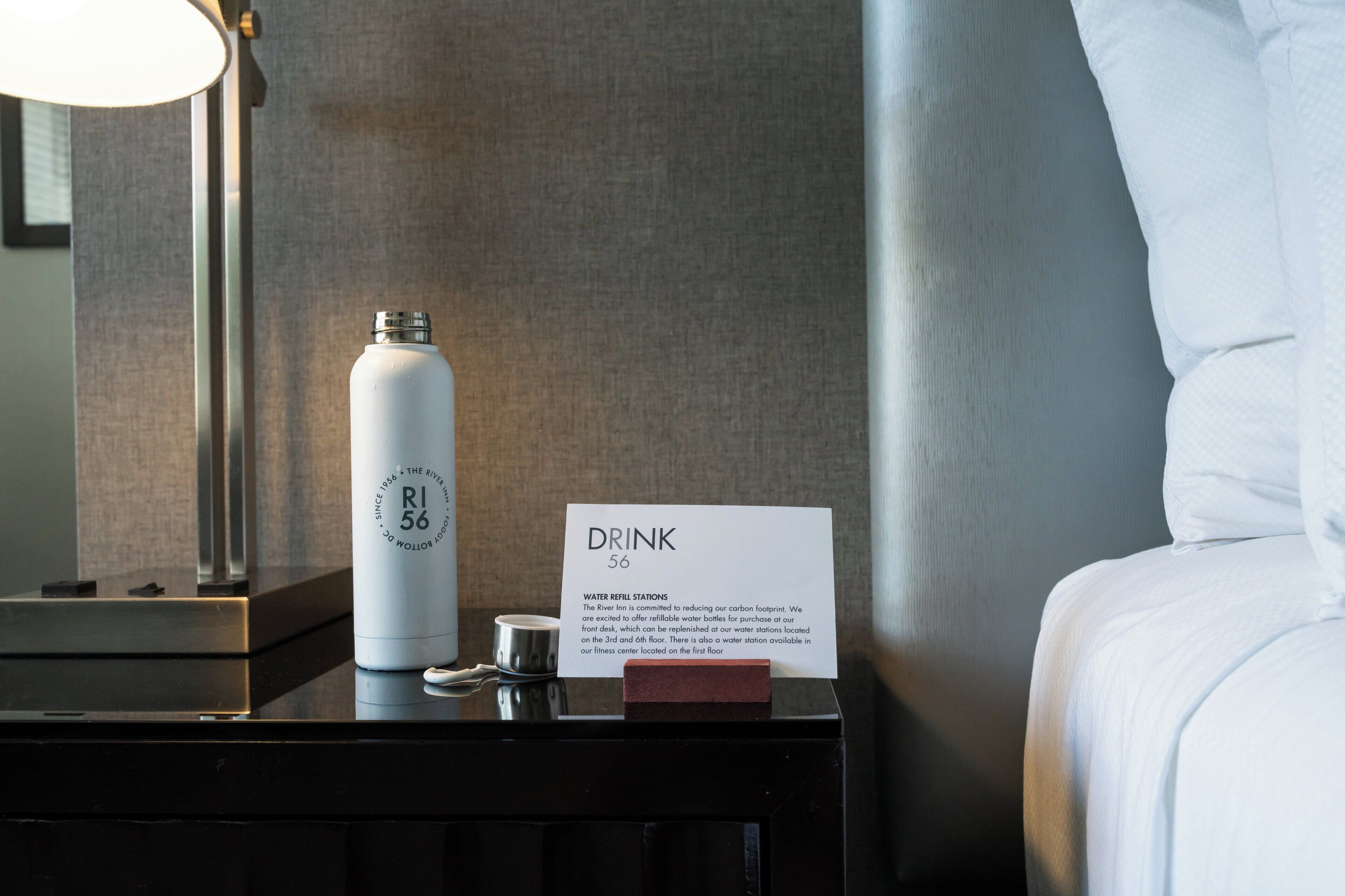 A hotel room with a water bottle on the nightstand, a sign indicating a drink refill station, and a lamp.