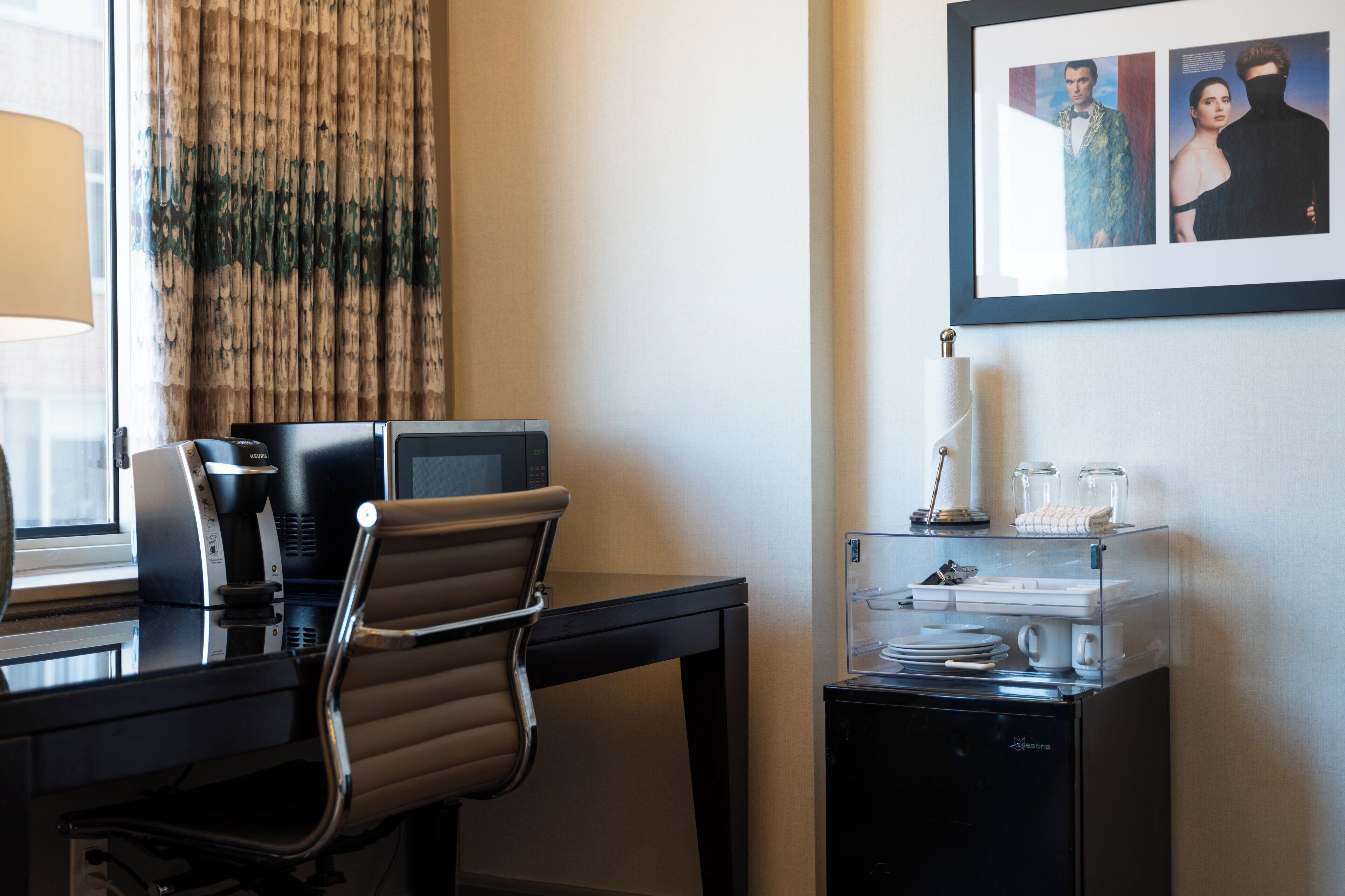 A hotel room with a desk, a chair, a coffee machine, a small refrigerator, and a framed picture on the wall.