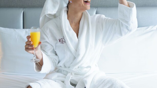 Una persona con bata blanca sentada en una cama con una bandeja de desayuno y un vaso de jugo de naranja.