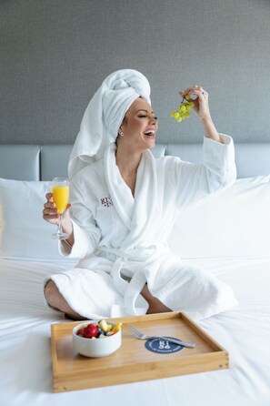 Una persona con bata blanca sentada en una cama con una bandeja de desayuno y un vaso de jugo de naranja.