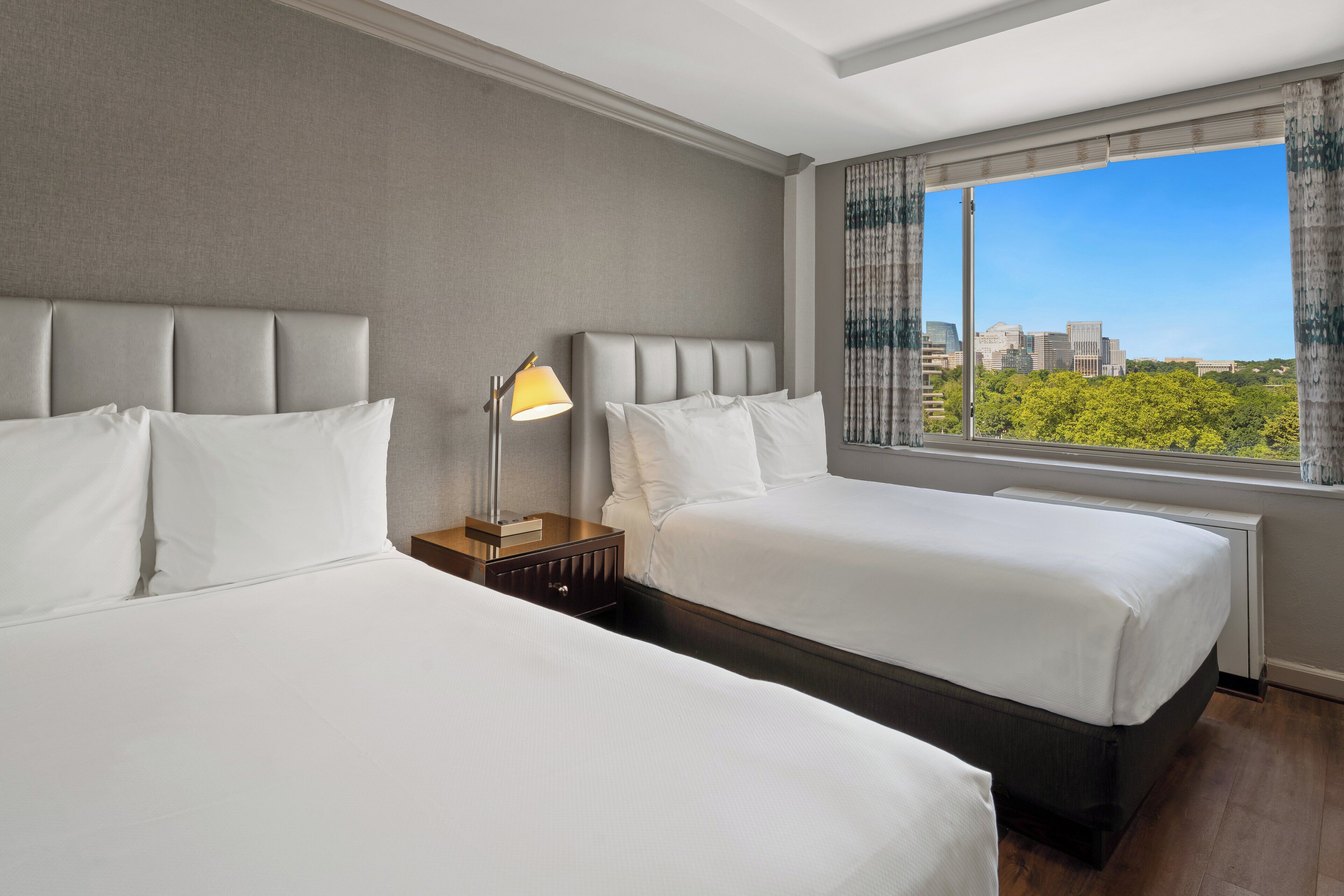 Habitación de hotel con dos camas, una mesita de noche, una lámpara y un ventanal con vistas a la ciudad.