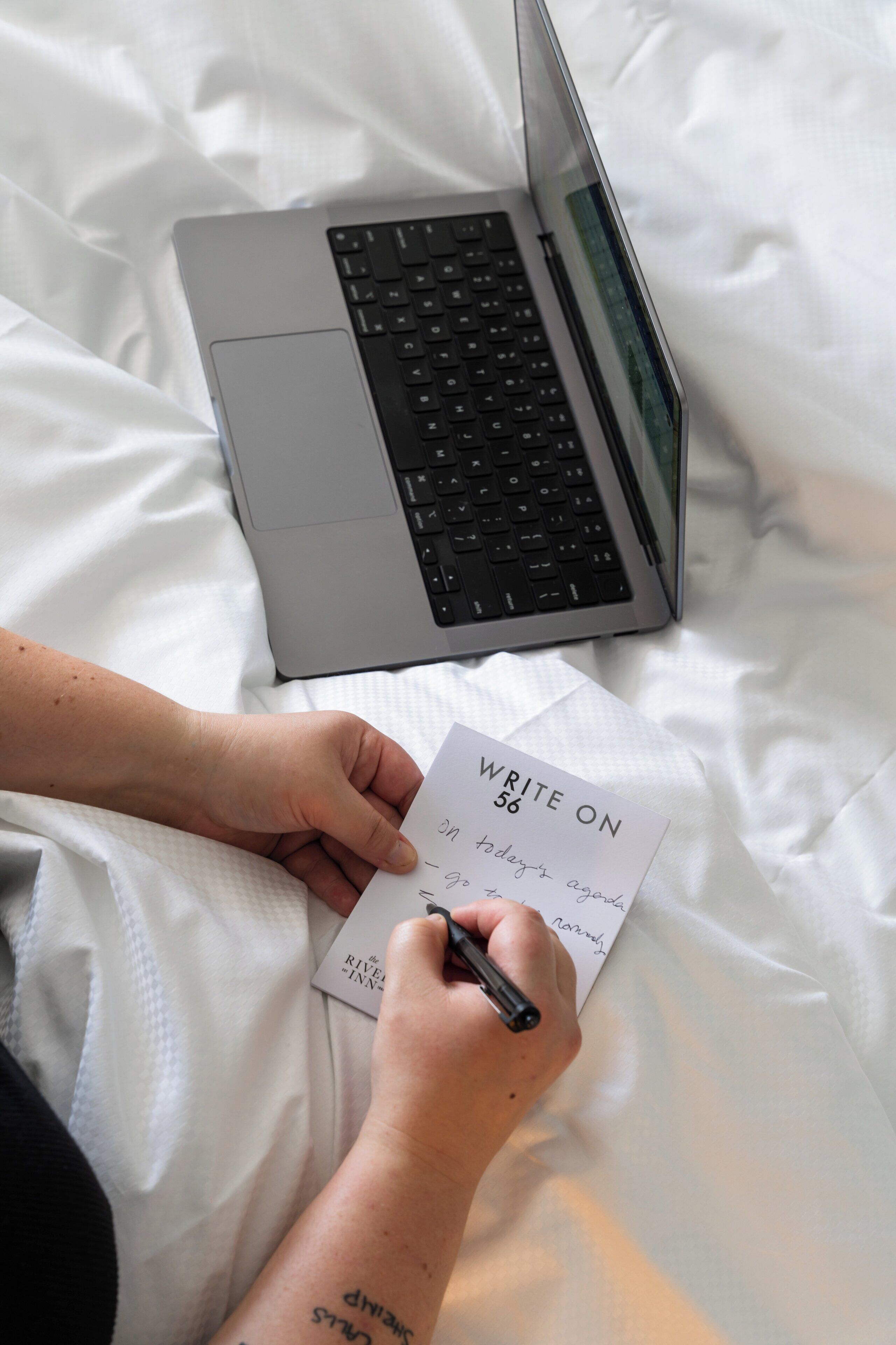 Um laptop numa cama, com um pedaço de papel com a inscrição 'WRITE ON' e uma caneta.