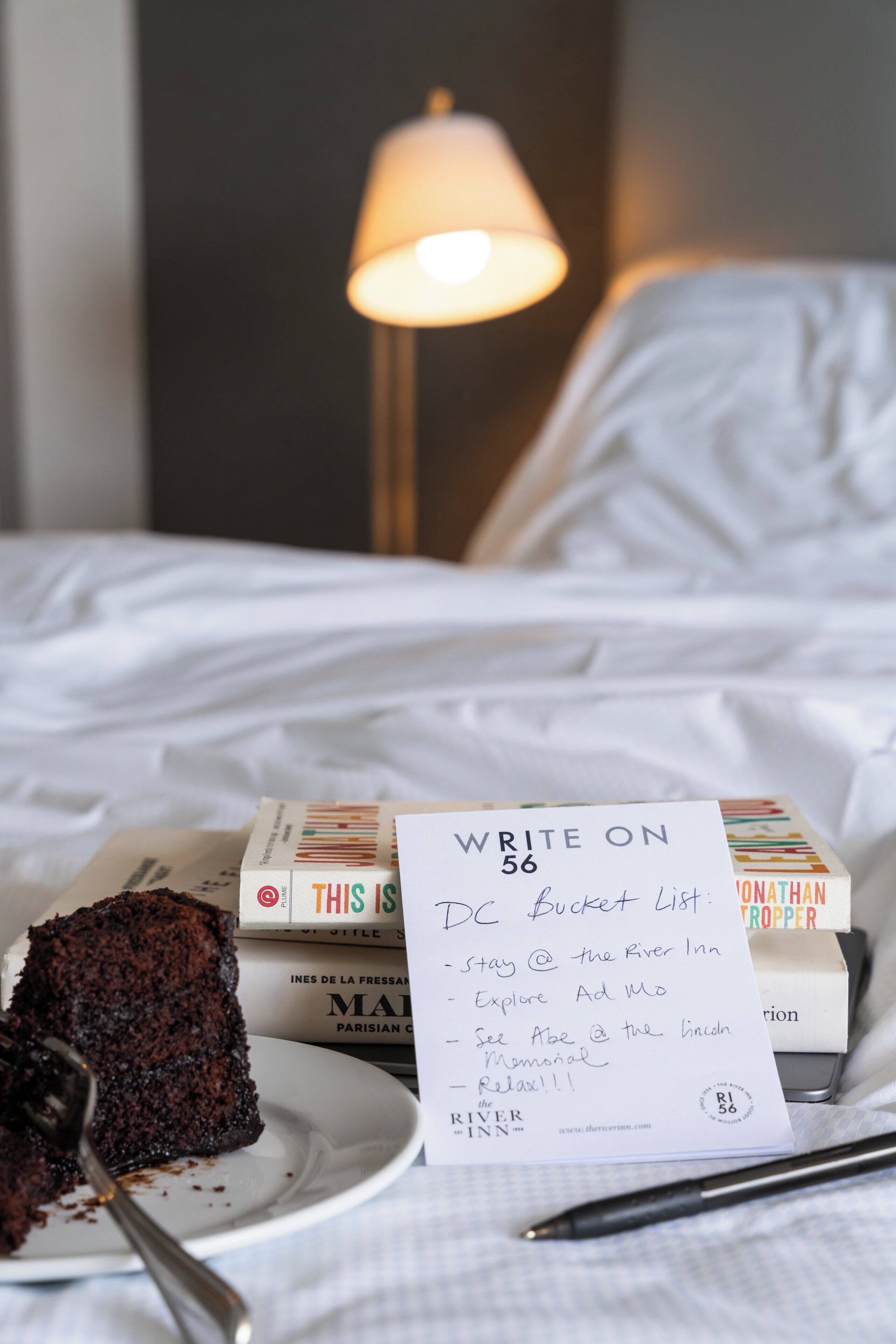 A bed with white linens, a bedside lamp, a plate with a piece of cake, a pen, and a note titled "WRITE ON DC Bucket List" with travel suggestions.