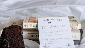 A bed with white linens, a bedside lamp, a plate with a piece of cake, a pen, and a note titled "WRITE ON DC Bucket List" with travel suggestions.
