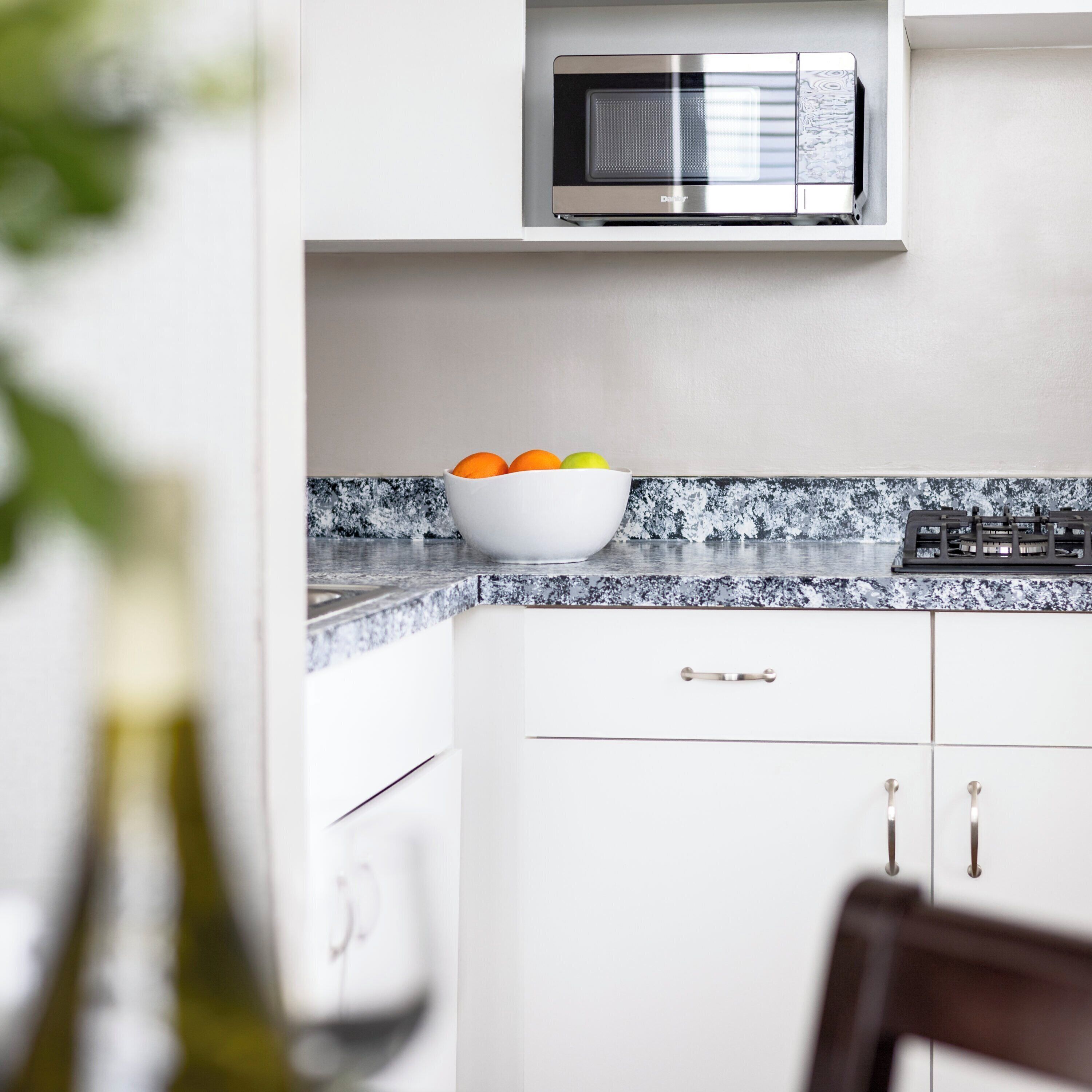 A kitchen with a white countertop, a bowl of fruit, a microwave, and a stove.