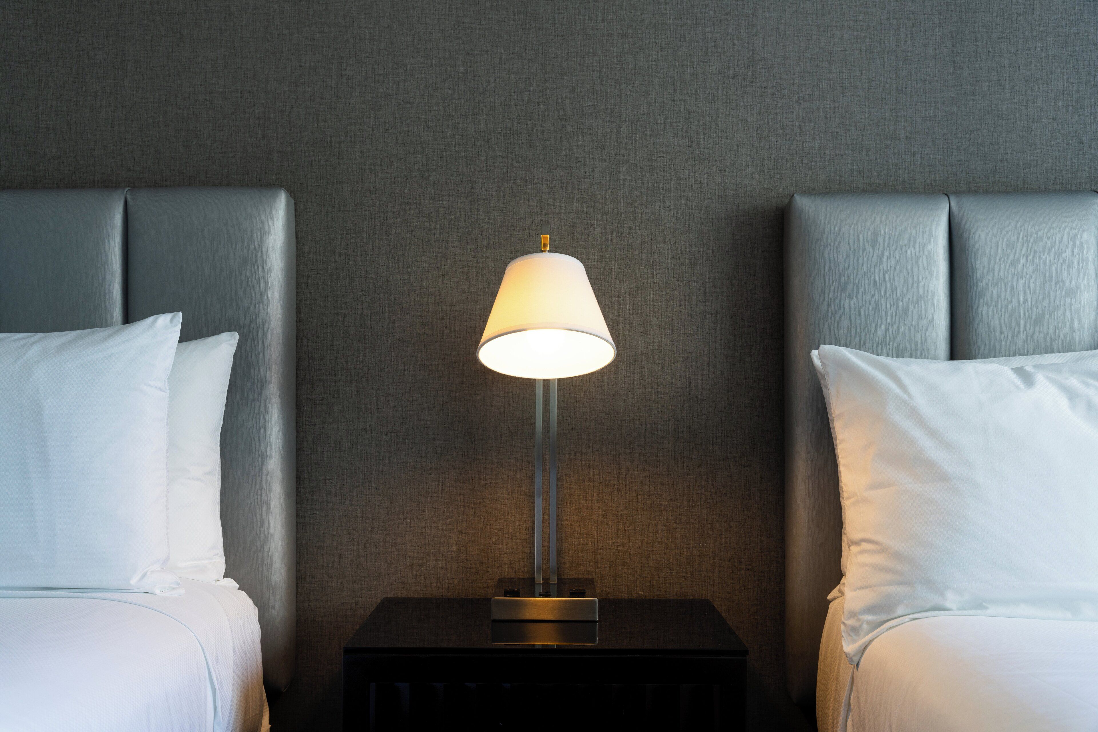Quarto de hotel com duas camas, uma mesa de cabeceira com abajur e parede texturizada.