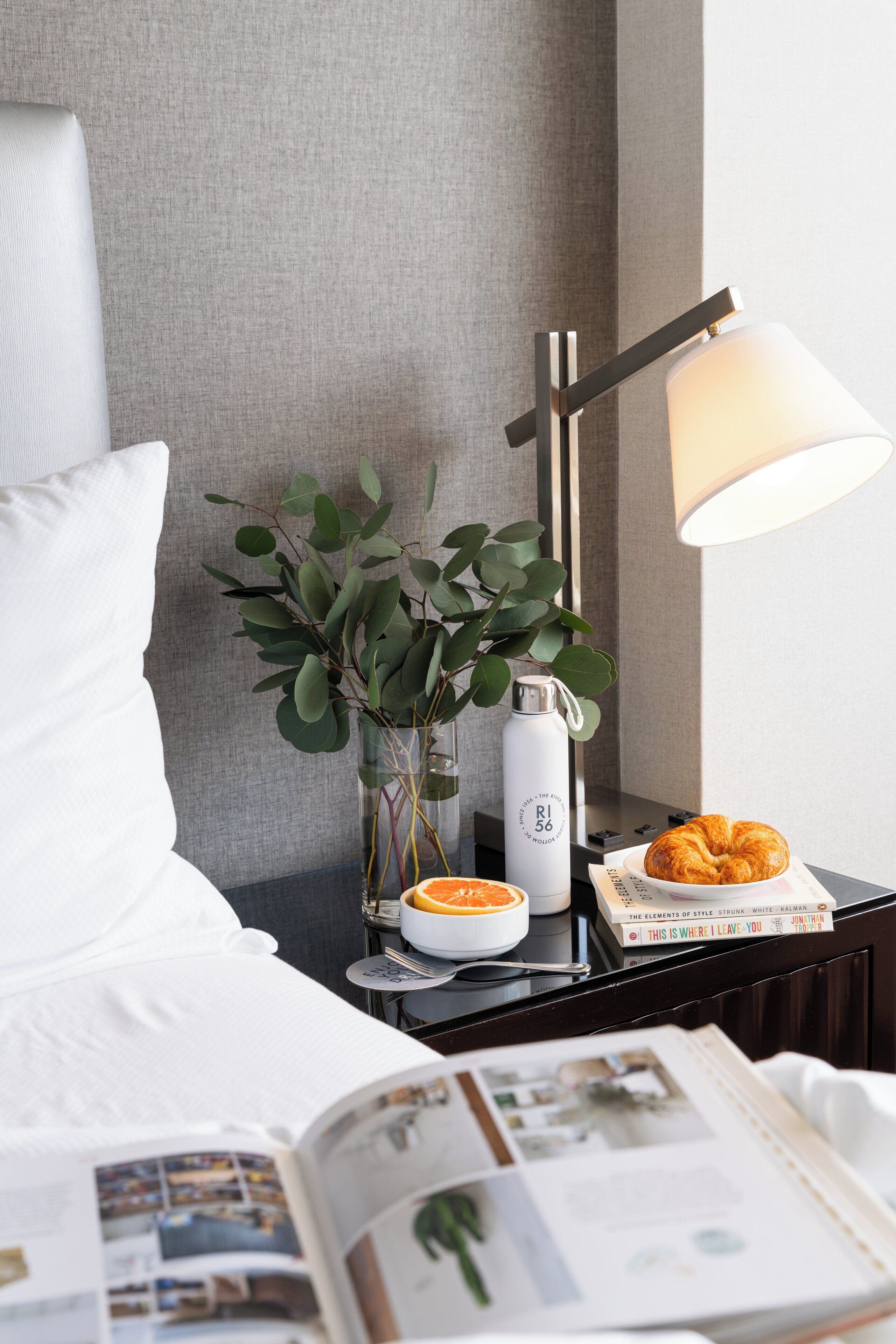 Ett välordnat hotellrum med en säng, ett sängbord med en lampa, en vas med gröna blad, en vattenflaska, en croissant och en bok med titeln ”HÄR LÄMNAR JAG DIG” av Jonathan White.
