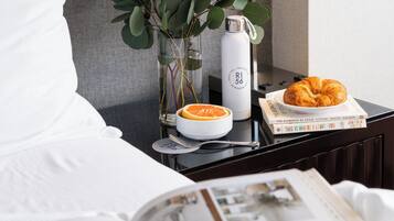 Ett välordnat hotellrum med en säng, ett sängbord med en lampa, en vas med gröna blad, en vattenflaska, en croissant och en bok med titeln ”HÄR LÄMNAR JAG DIG” av Jonathan White.