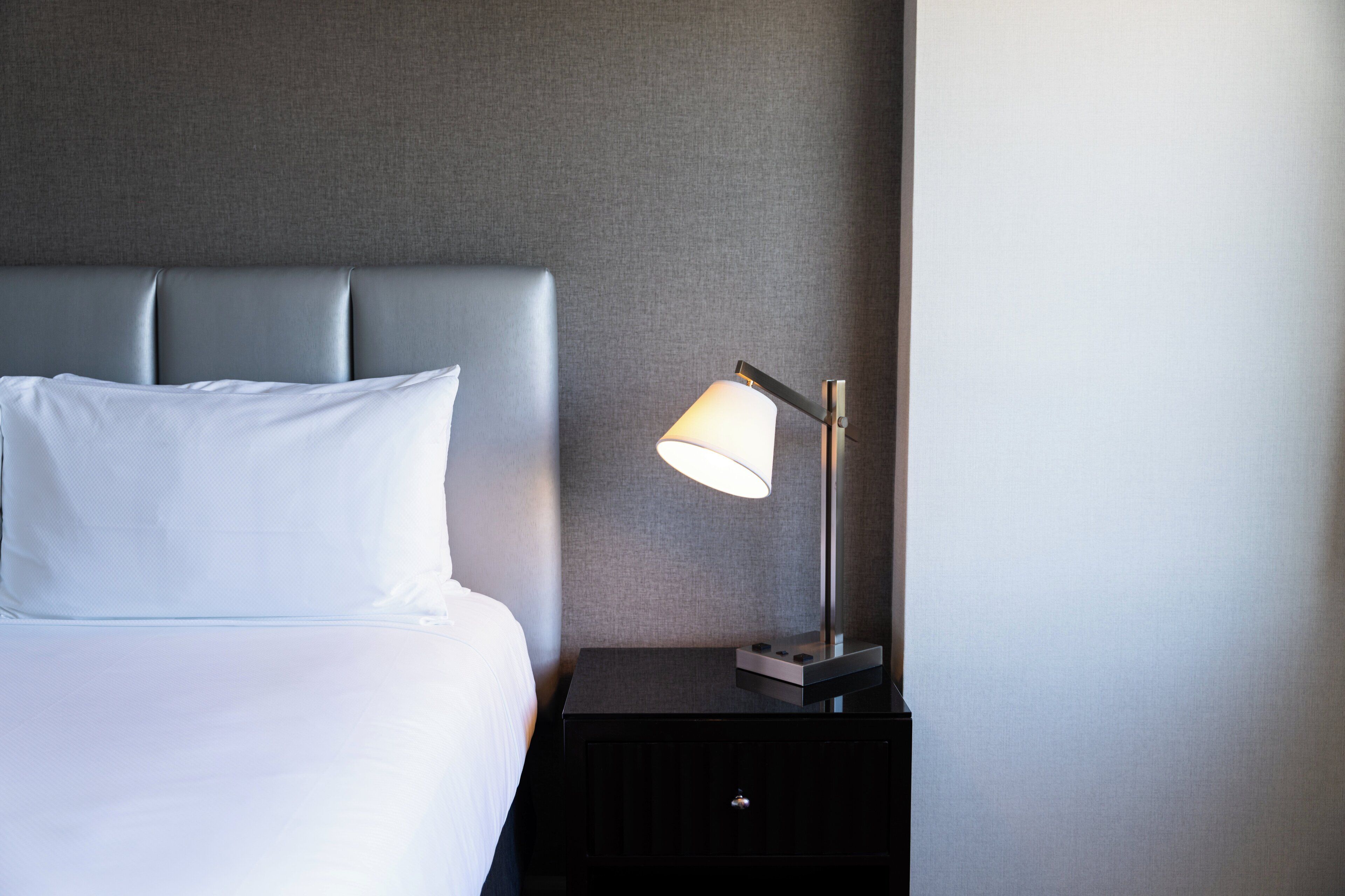 Una cama bien tendida con cabecera gris, una mesita de noche con lámpara y una pared gris.